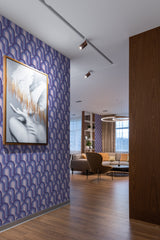 The wallpaper has a purple background with a repeating pattern of curved lines and arches in shades of purple, white, and gray, reminiscent of retro design. A framed artwork with abstract imagery hangs on the wall. Wood flooring and soft lighting complement the room's aesthetic, creating a cozy atmosphere with traditional wallpaper.