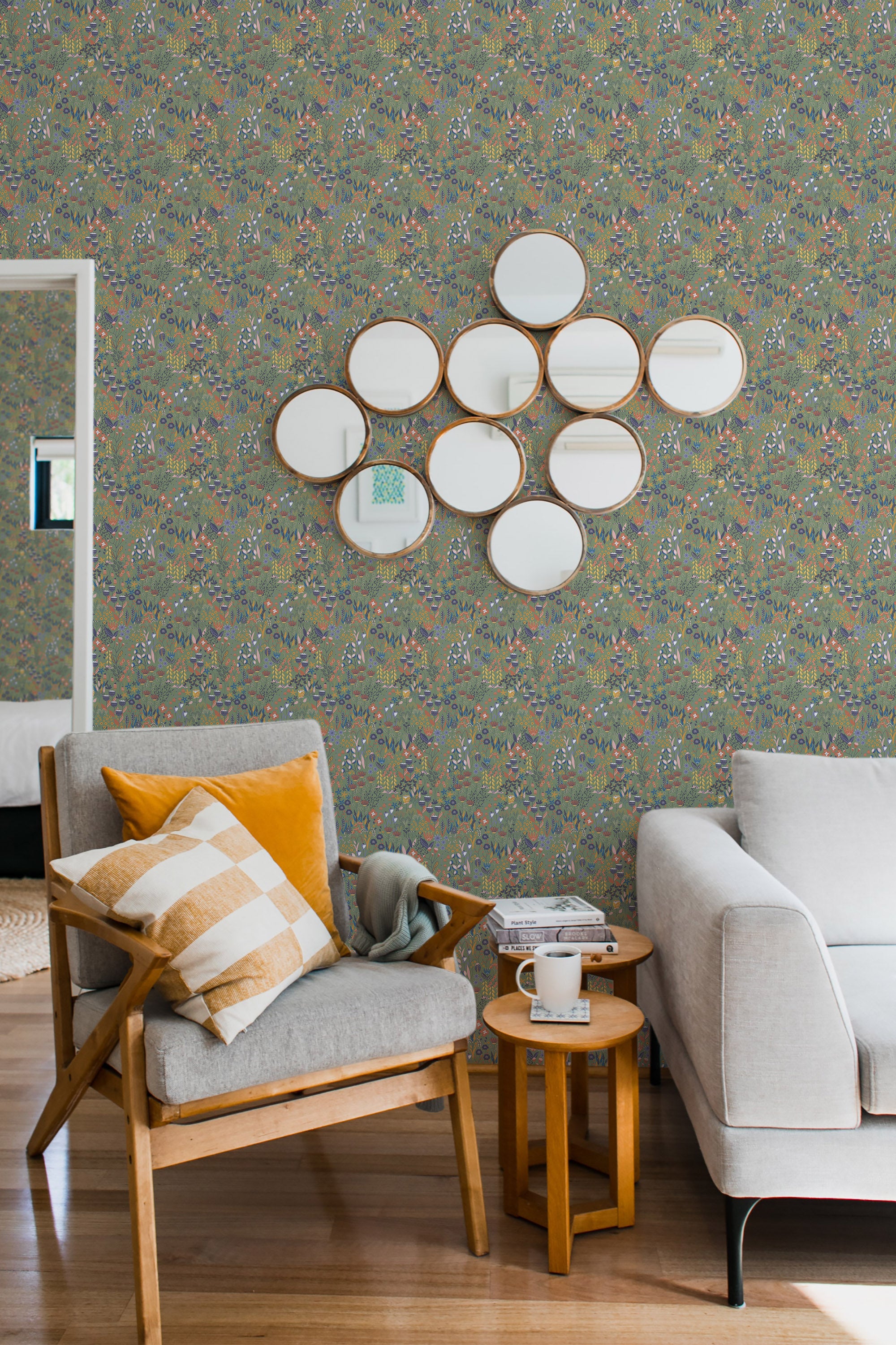 A cozy room with a wall covered in Sage Meadow wallpaper, a pattern with delicate flowers and leaves in muted colors, complemented by round mirrors and modern furniture, invoking a serene ambiance with traditional wallpaper.