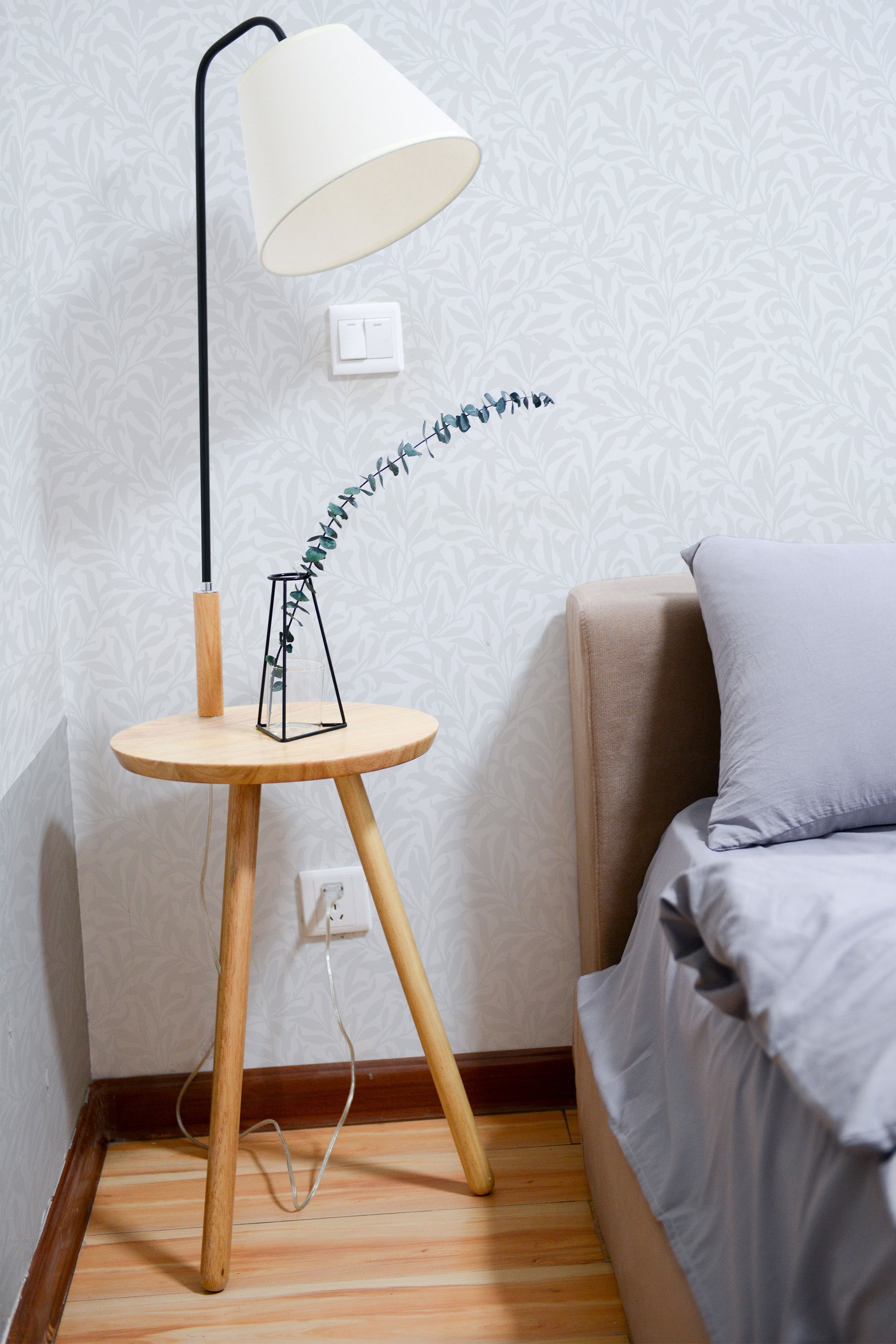 Self-adhesive wallpaper with seamless spring leaves pattern, light pastel tones, in a cozy room with a bedside table and lamp.
