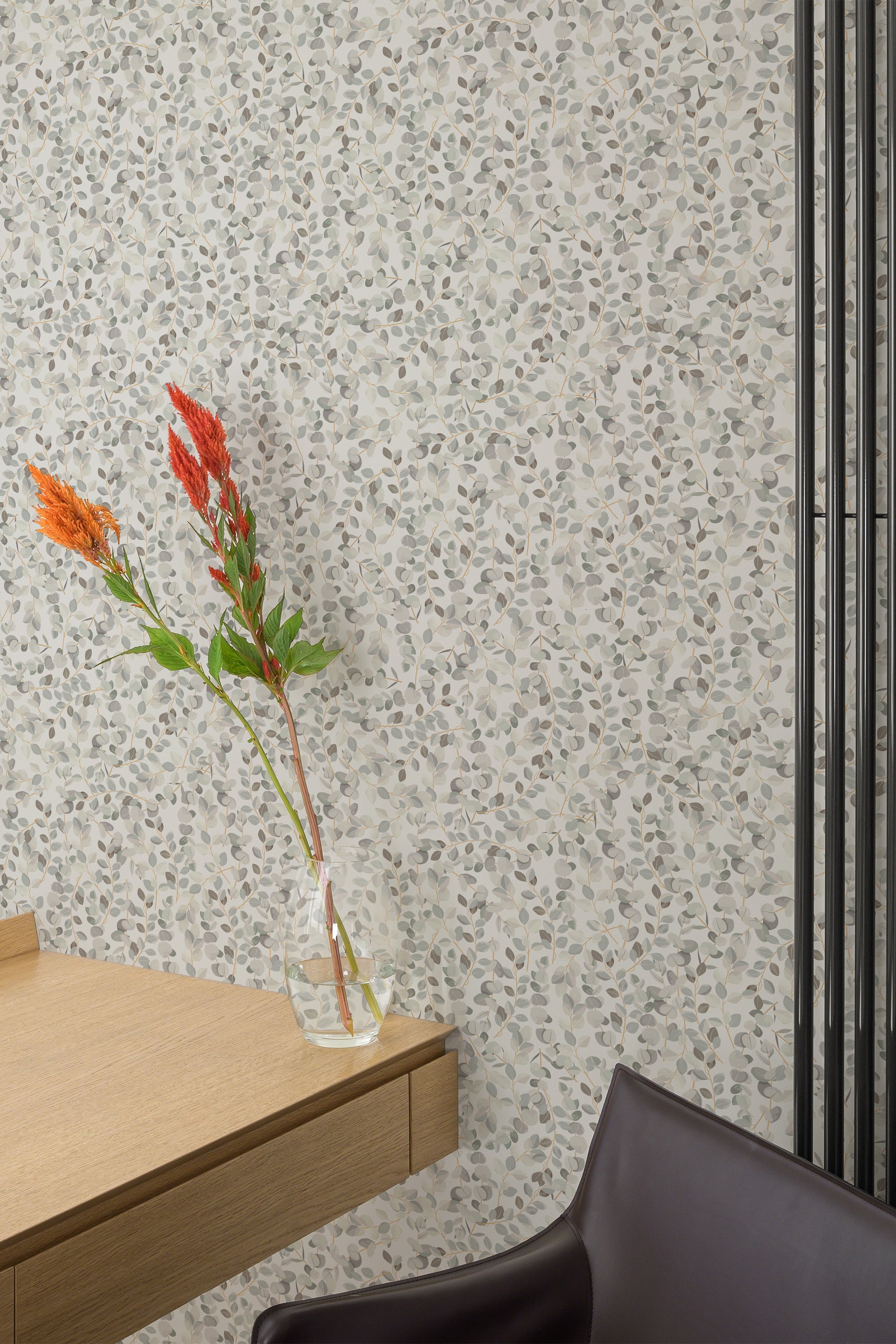 Self-adhesive wallpaper with a pattern of aesthetic spring leaves in various shades of gray adorning a room's wall behind a wooden table and a clear vase with red flowers.
