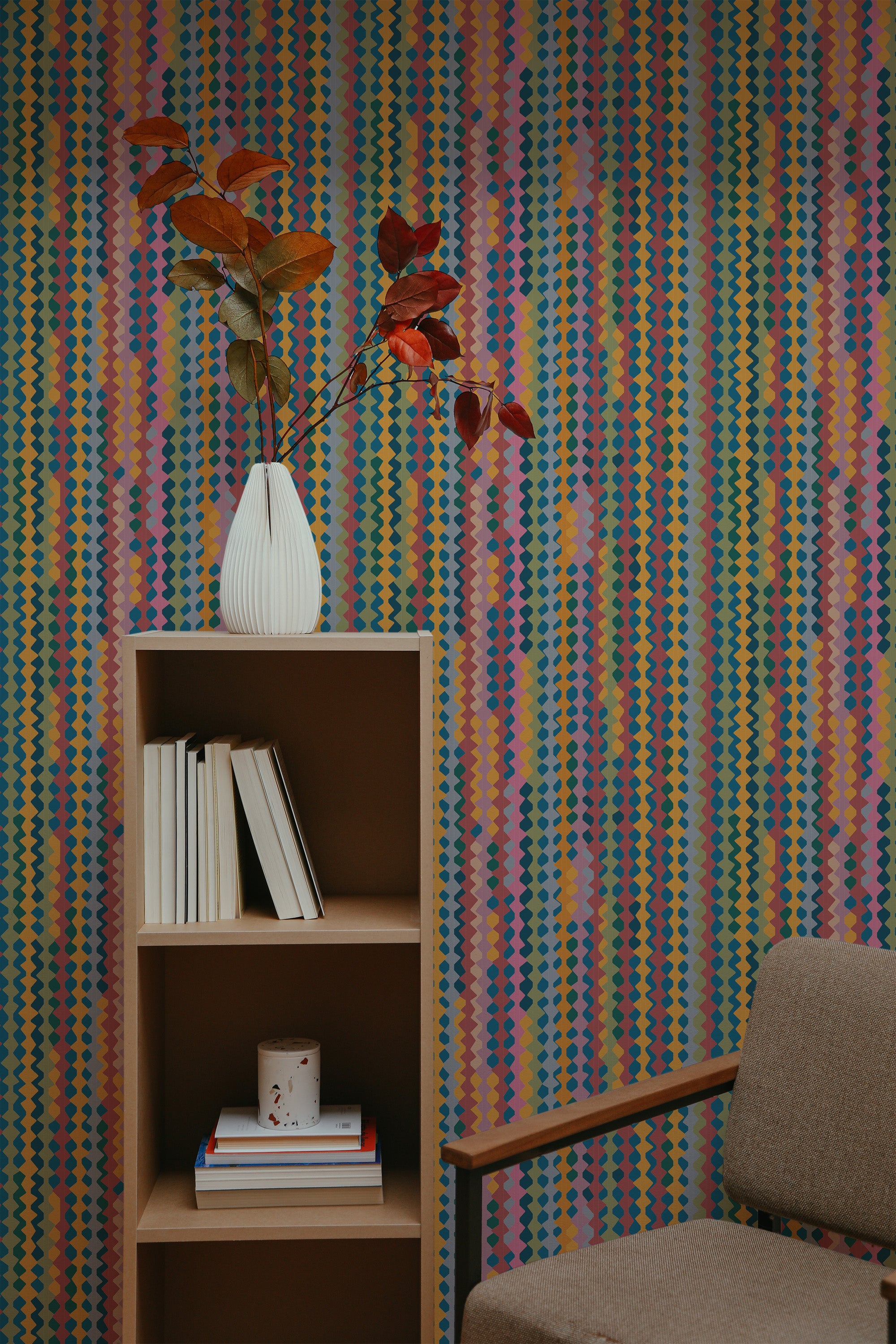 Self-adhesive retro vintage geometric wallpaper with colorful vertical stripes and shapes on a room wall, complemented by a wooden bookshelf and a gray armchair.