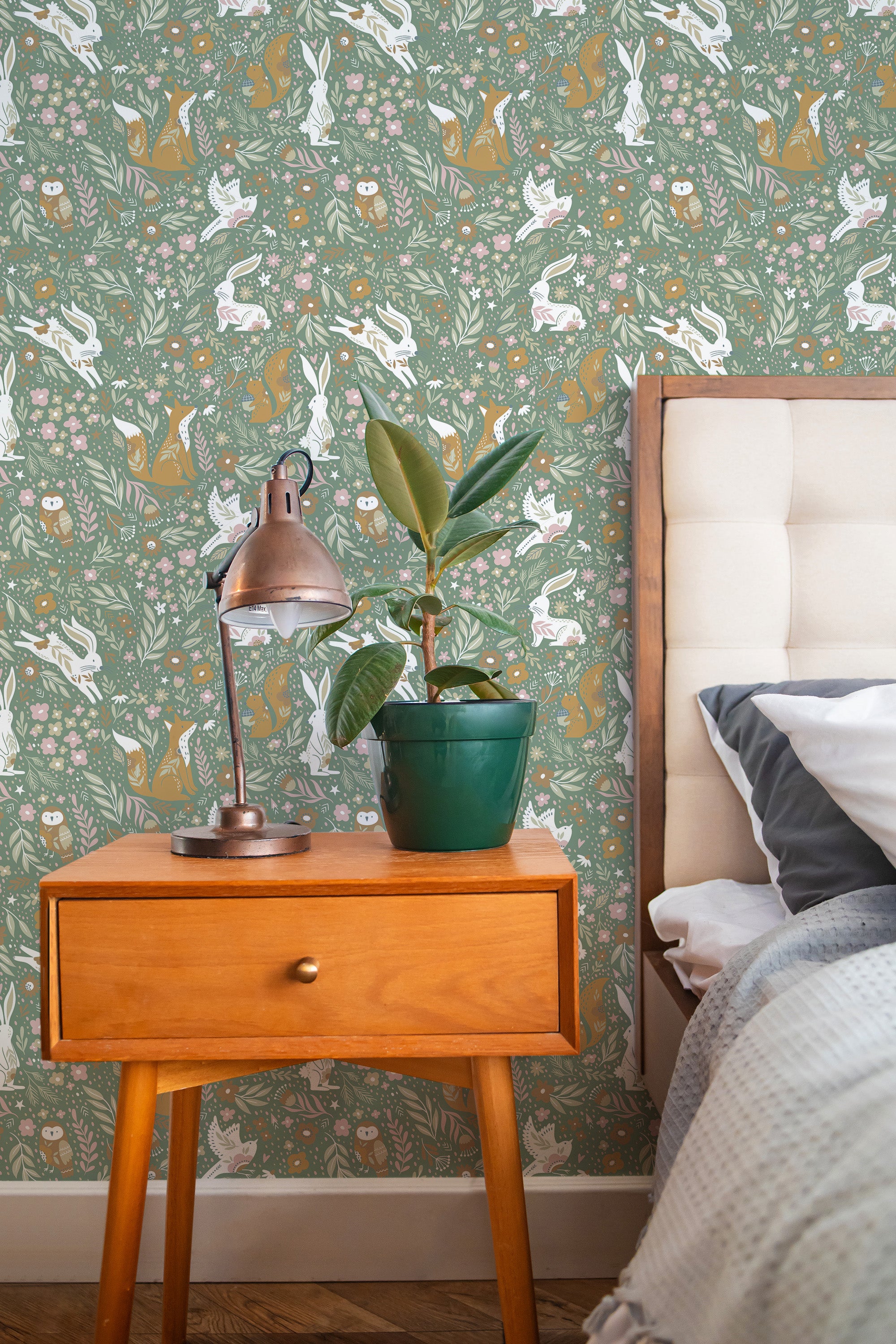 Self-adhesive Sage magical forest wallpaper with rabbits, birds, and trees on a bedroom wall, accompanied by a wooden bedside table, lamp, and potted plant.