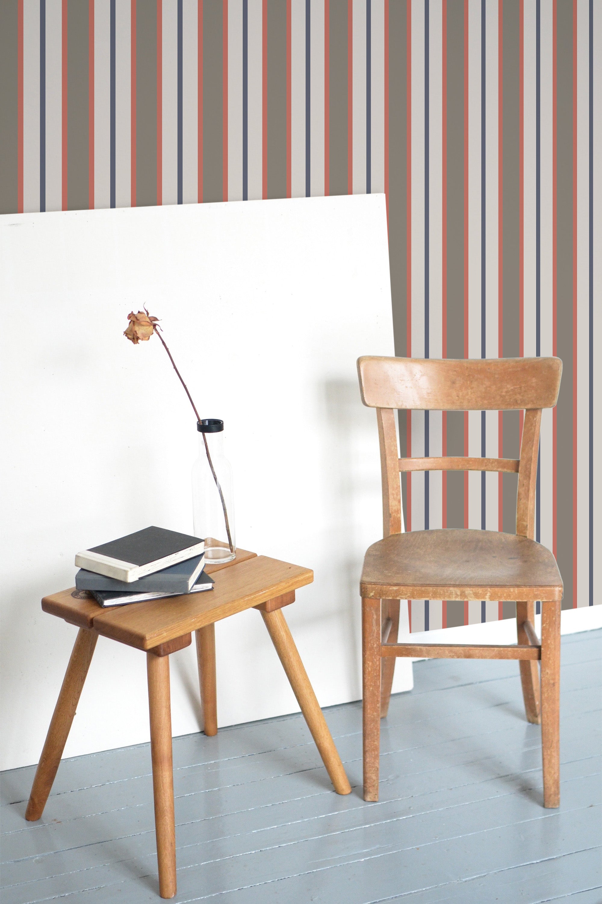 Self-adhesive wallpaper with vertical earthy-toned stripes in varying widths on a wall, with a wooden chair and side table in the foreground.