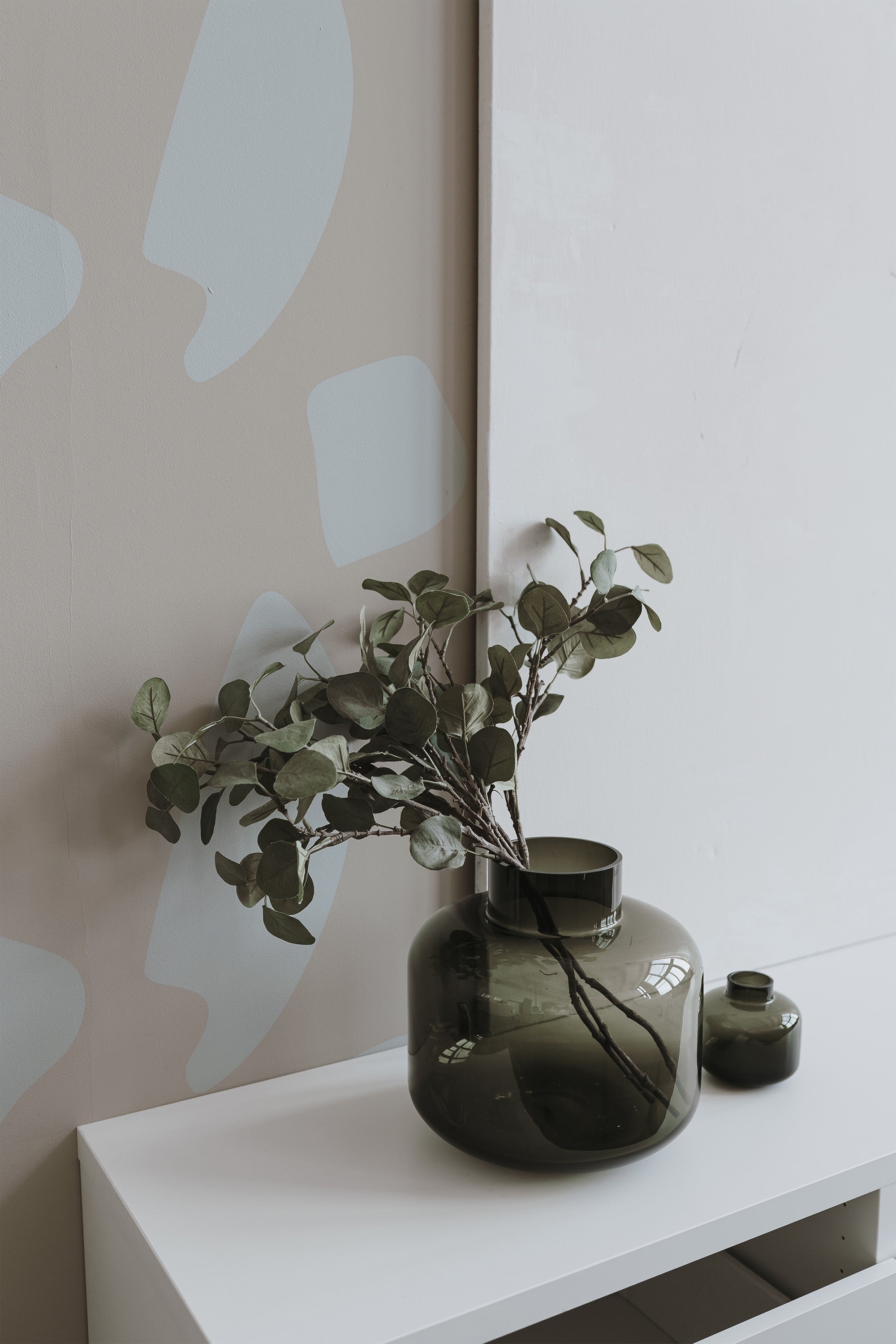 Self-adhesive wallpaper with abstract neutral-toned geometric shapes on a wall, complemented by a vase with greenery on a white shelf.