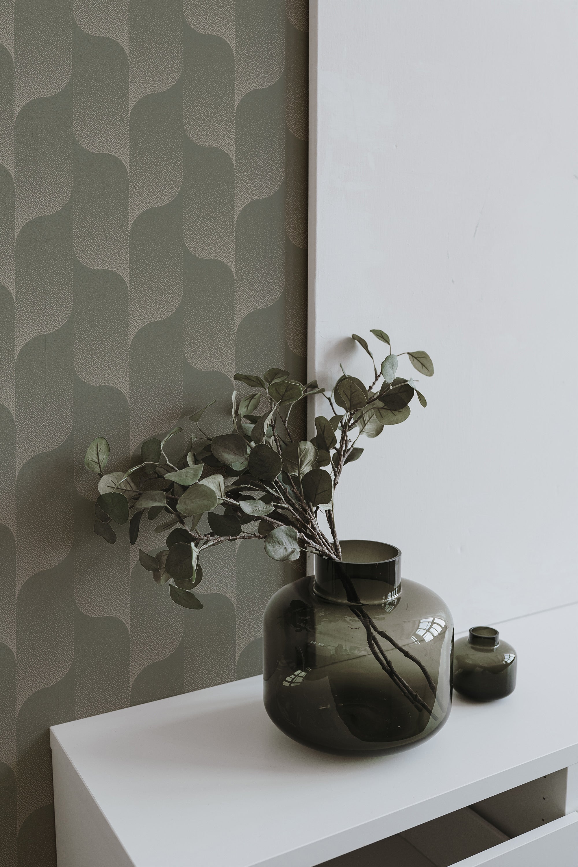 Self-adhesive wallpaper with a smooth gradient of green hues in a wavy pattern on a room wall, complemented by a vase with greenery on a white shelf.