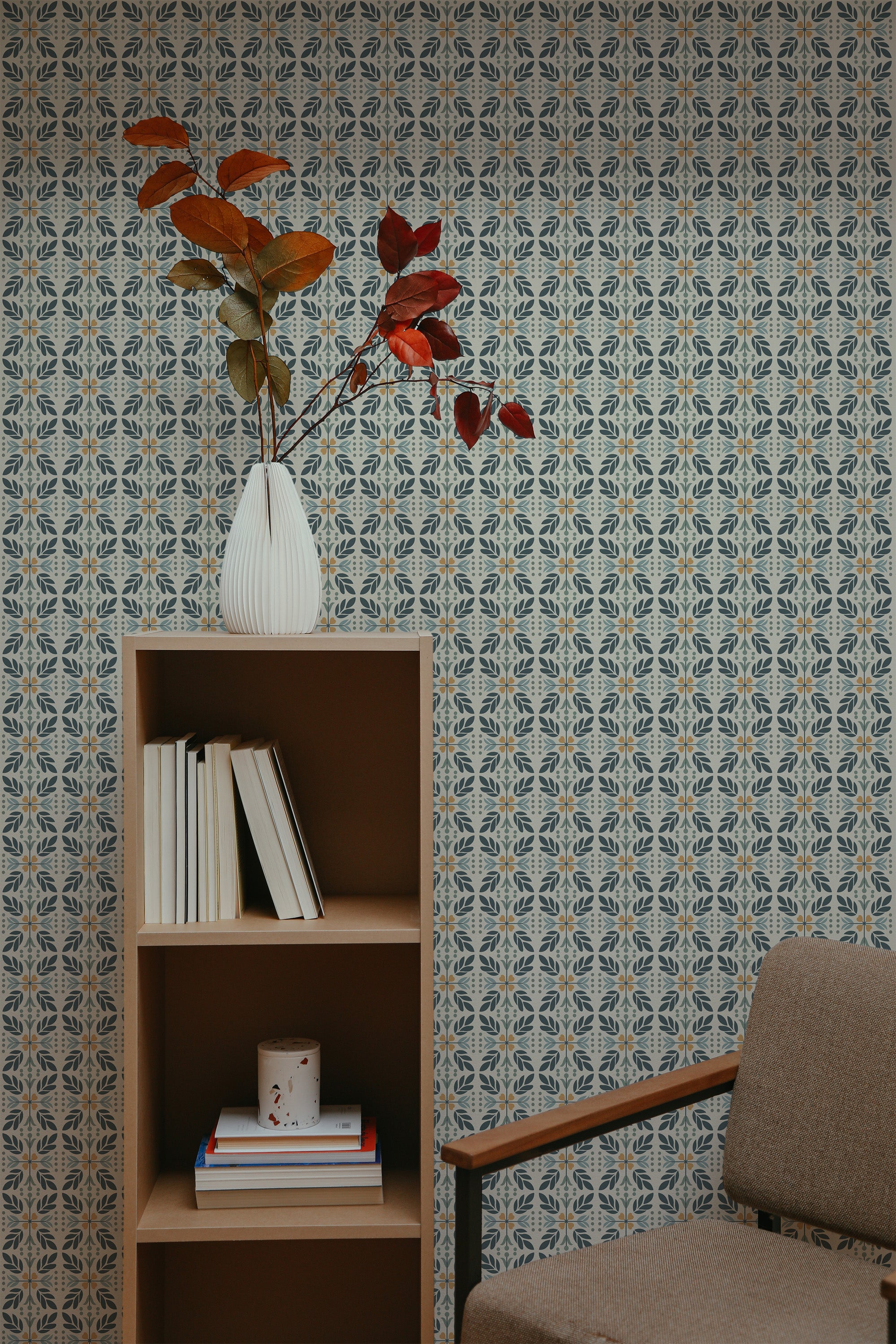Self-adhesive wallpaper with intricate busy tile pattern in shades of blue and beige, covering a wall in a room with a wooden bookshelf, a vase with red leaves, and a brown armchair.