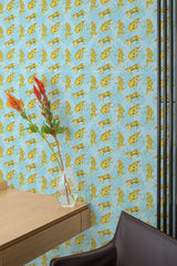 Self-adhesive wallpaper with a bold pattern of yellow tigers on a blue background in a room with a wooden desk and a vase with flowers.