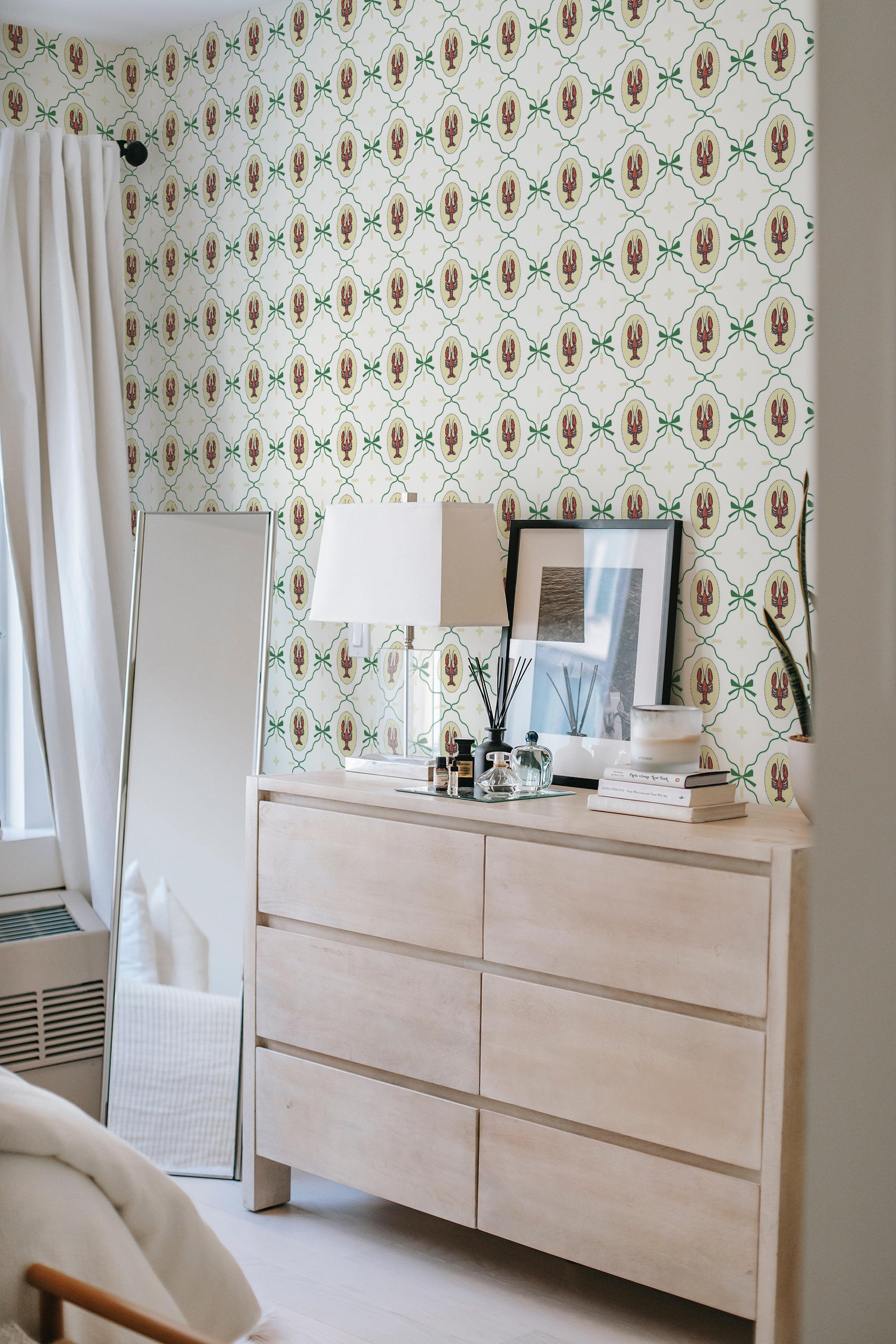 A bedroom with a dresser in the foreground and walls covered in a repeating pattern of red lobsters surrounded by green foliage on a cream background, traditional wallpaper.