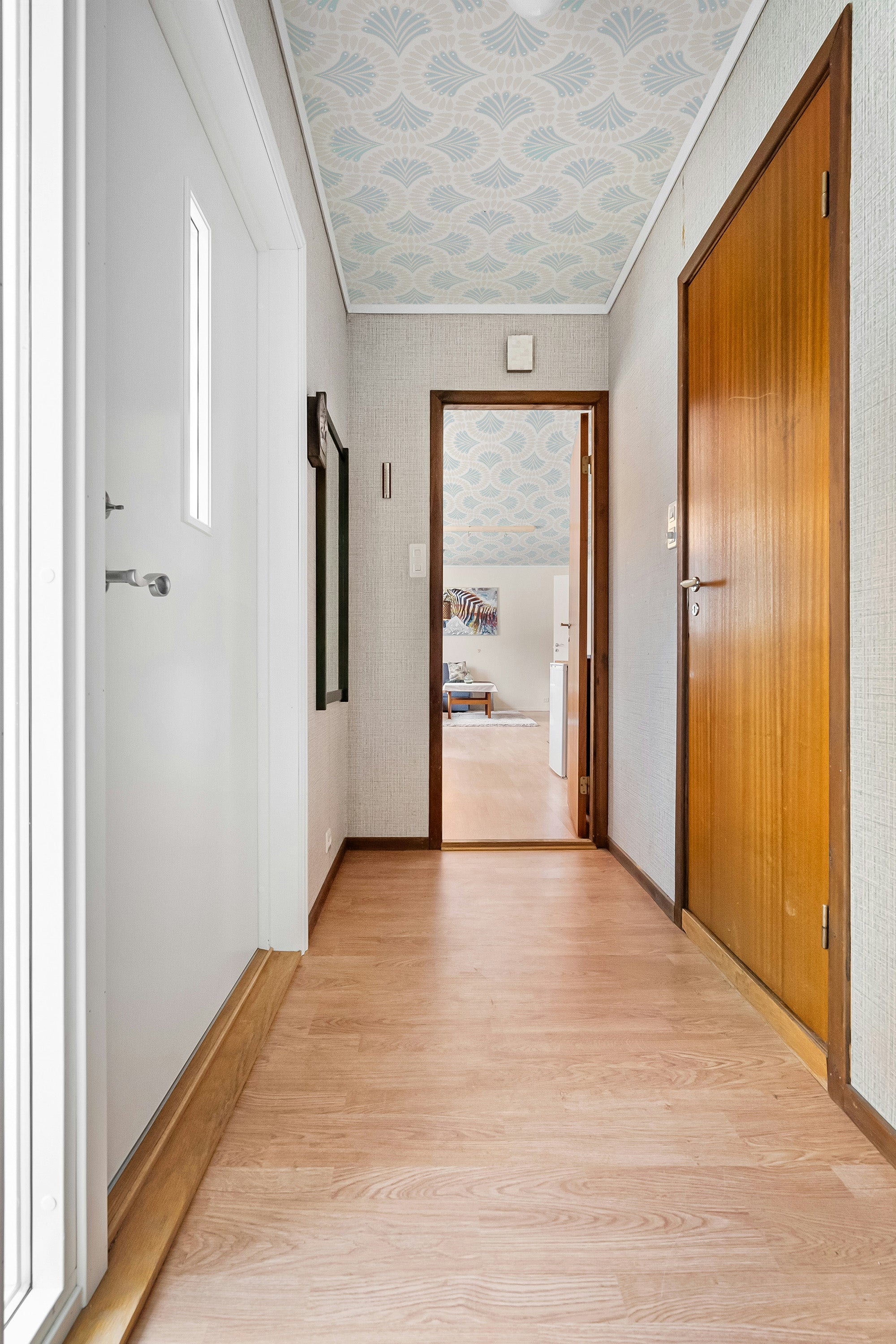 Hallway with wooden floor, white walls, and a wooden door and ceiling wallpaper.
