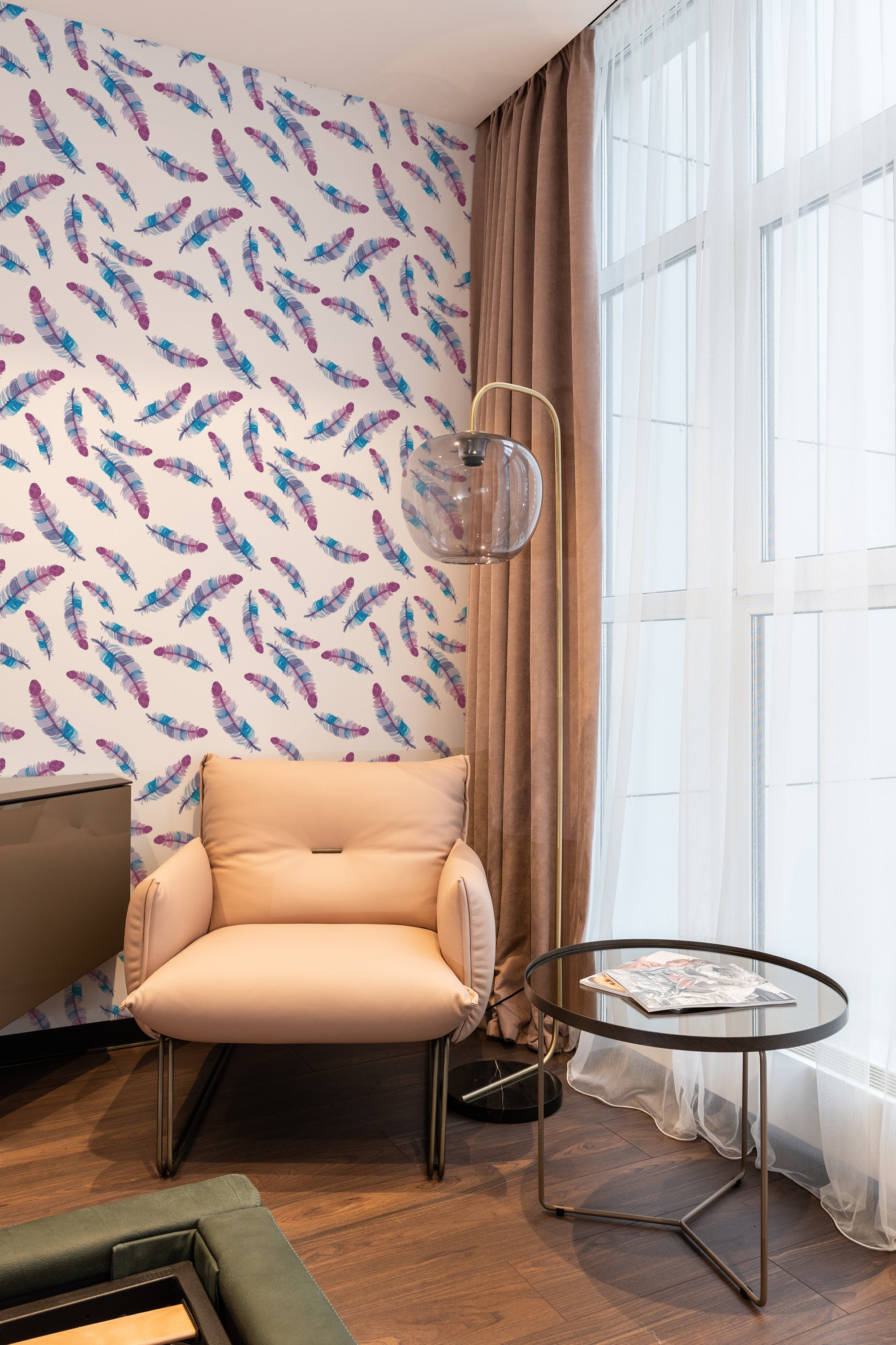 A room with wallpaper adorned with delicate, multicolored watercolor feathers in soft shades of blue, purple, and pink scattered across a light background. The room has a cozy armchair, a side table, and floor-length curtains near a large window. Traditional wallpaper.