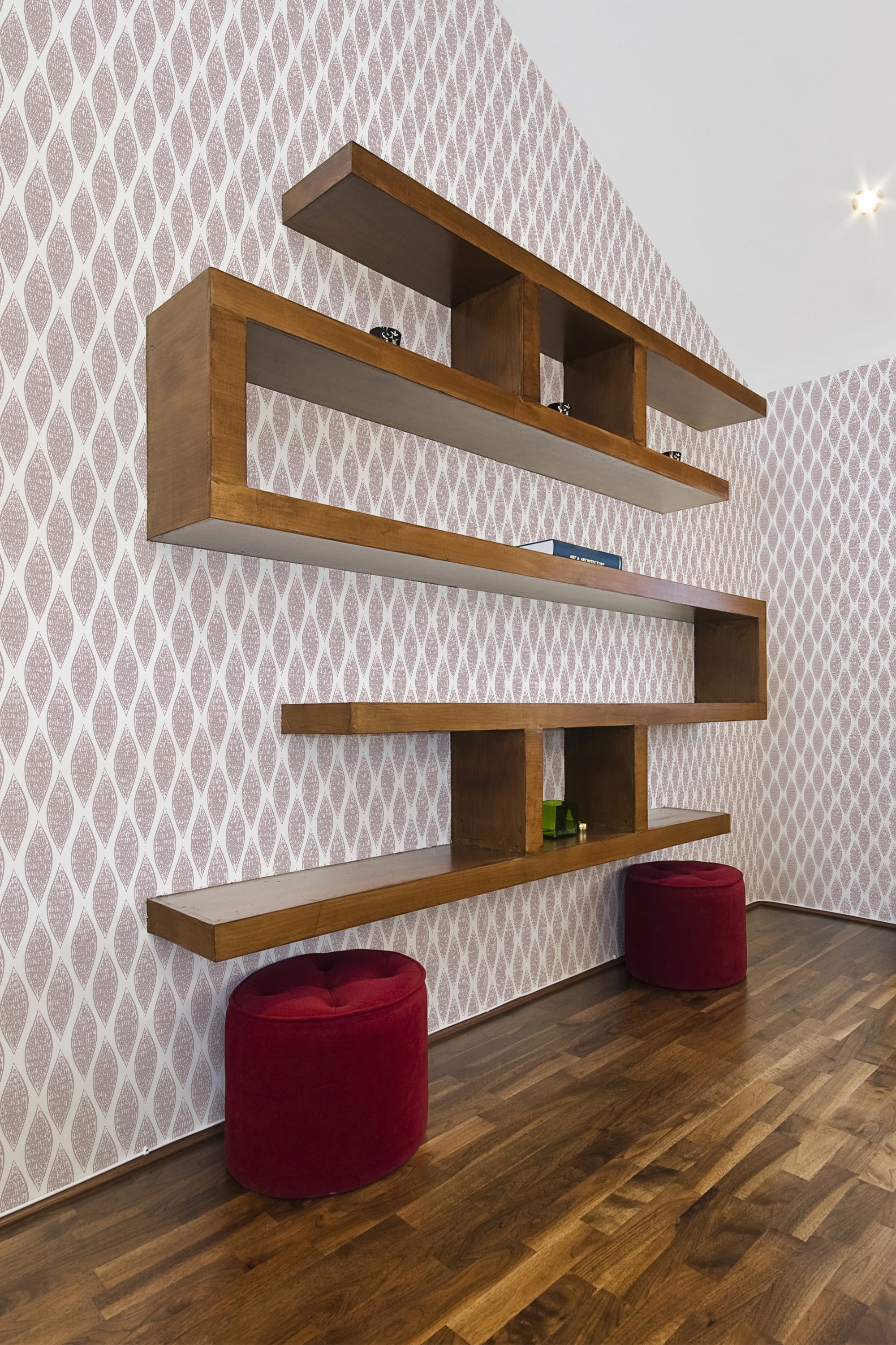 The wallpaper design includes repeated patterns of delicate, overlapping leaves in soft, subtle hues, creating an elegant and understated backdrop, complementing the wooden shelves and red stools in the room. Traditional wallpaper.