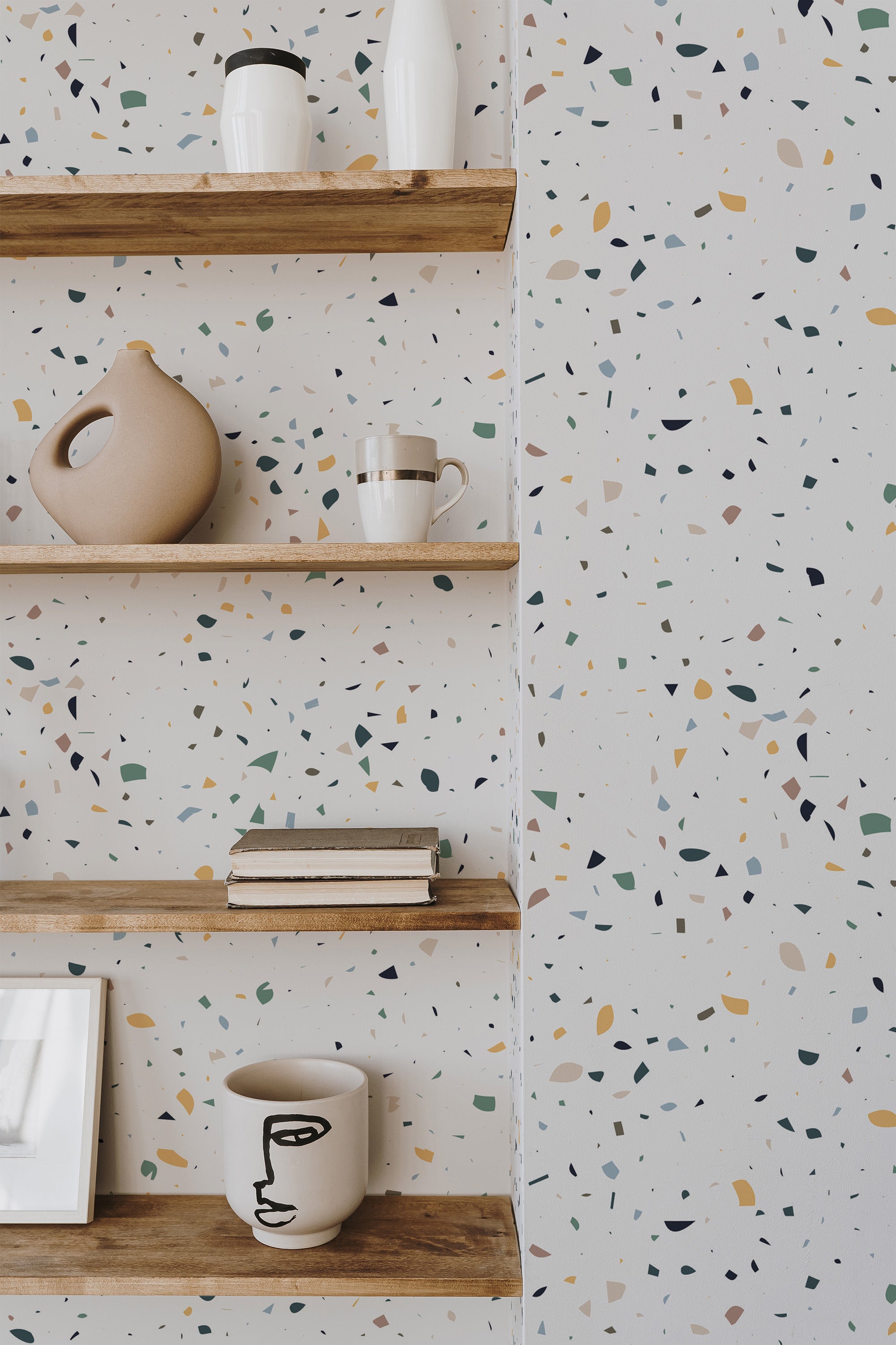 Self-adhesive wallpaper with a Terrazzo floor texture displaying an irregular scattering of multi-colored speckles in earthy tones, including beige, olive, and ochre, against a light background.