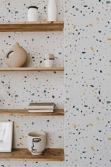 Self-adhesive wallpaper with a Terrazzo floor texture displaying an irregular scattering of multi-colored speckles in earthy tones, including beige, olive, and ochre, against a light background.