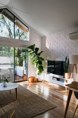 This image shows a light-filled room with walls covered in a Pink Coral design. The wallpaper has a soft pink coral pattern set against a light background, adding a touch of natural elegance. The room is decorated with minimalistic furniture, a large potted plant, and wooden flooring, creating a cozy yet modern atmosphere. Traditional wallpaper.