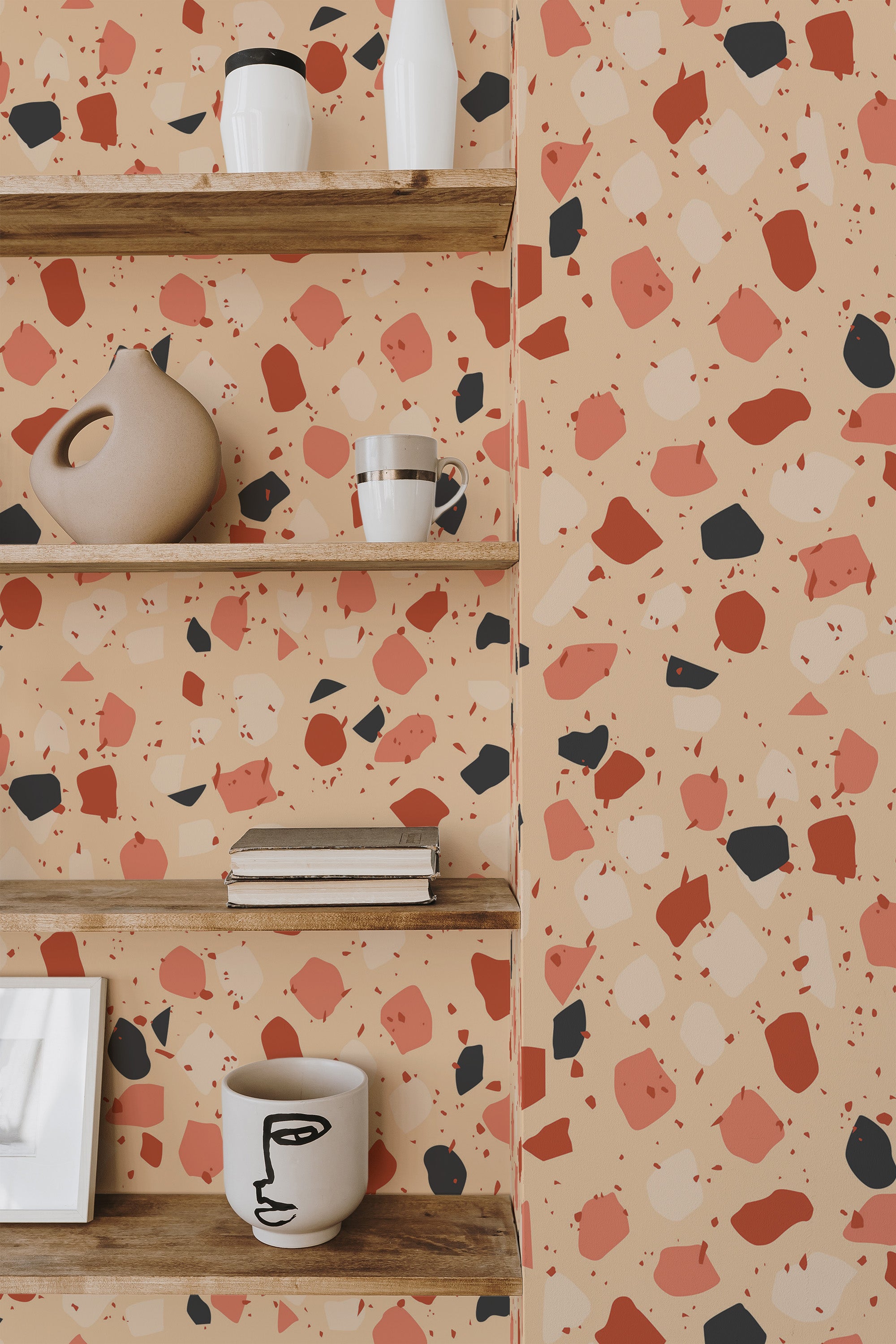 Self-adhesive wallpaper with a terrazzo design in a mix of peach, pink, black, and cream-colored irregular shapes on a beige background, covering the wall behind wooden shelves holding modern ceramic decor.