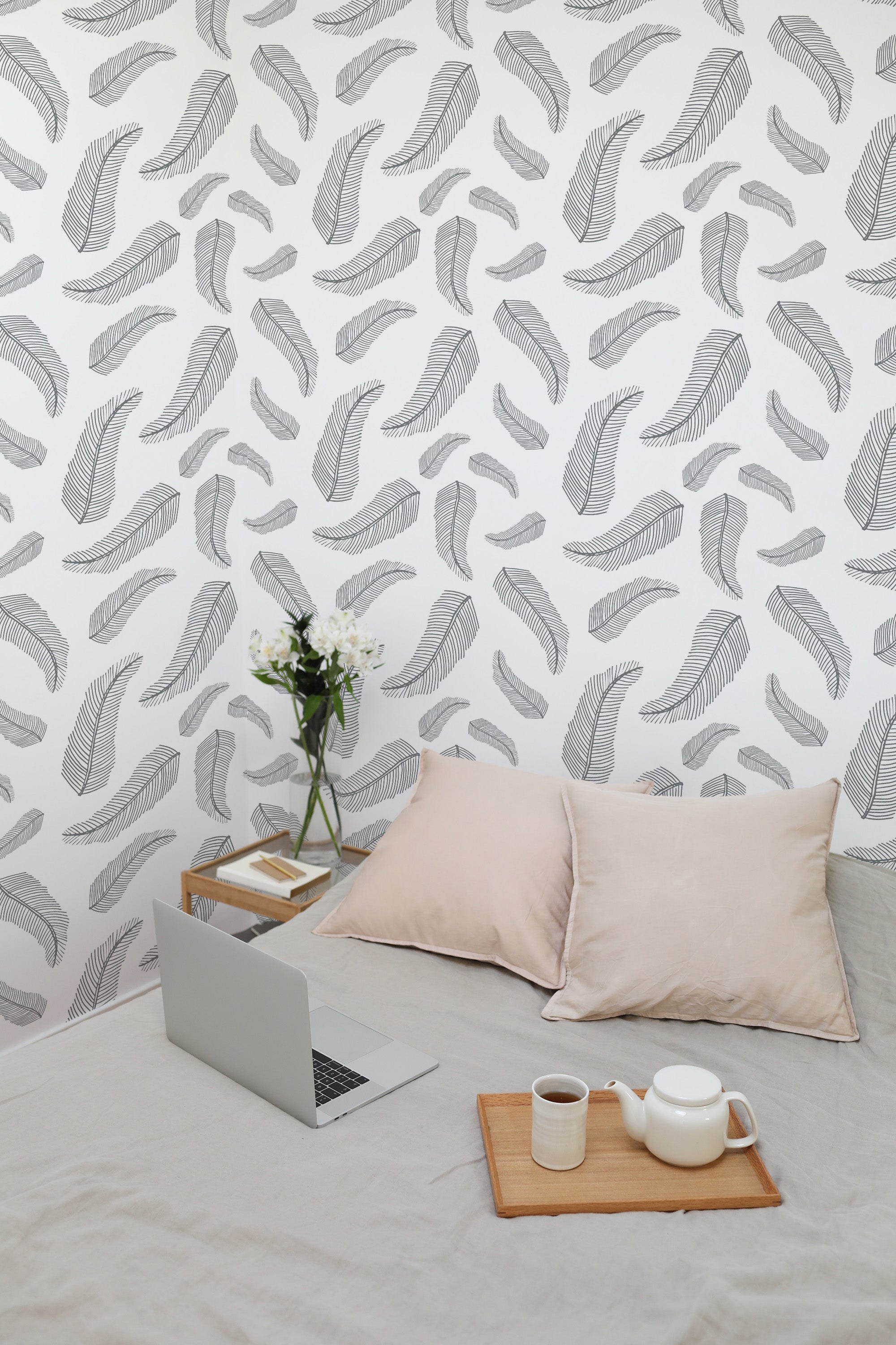 Self-adhesive wallpaper with a feather design in soft, neutral tones decorating the wall behind a bed with light-colored pillows, a small wooden tray holding a teapot and cup, and a vase of white flowers on a side table.