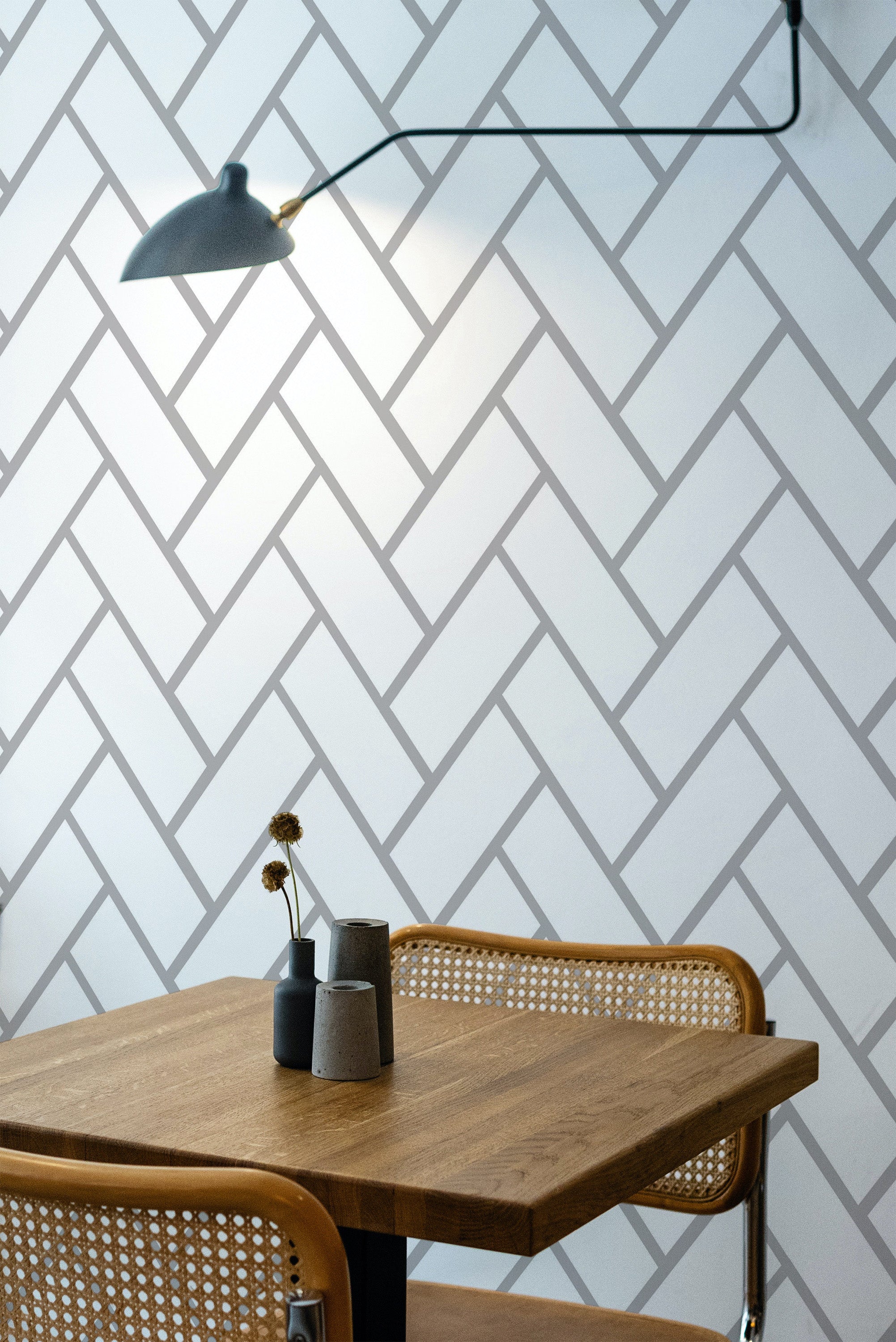 Self-adhesive herringbone tile wallpaper with a chevron pattern in shades of white and light gray, covering the wall behind a wooden table and wicker chairs, with a modern black wall lamp overhead.