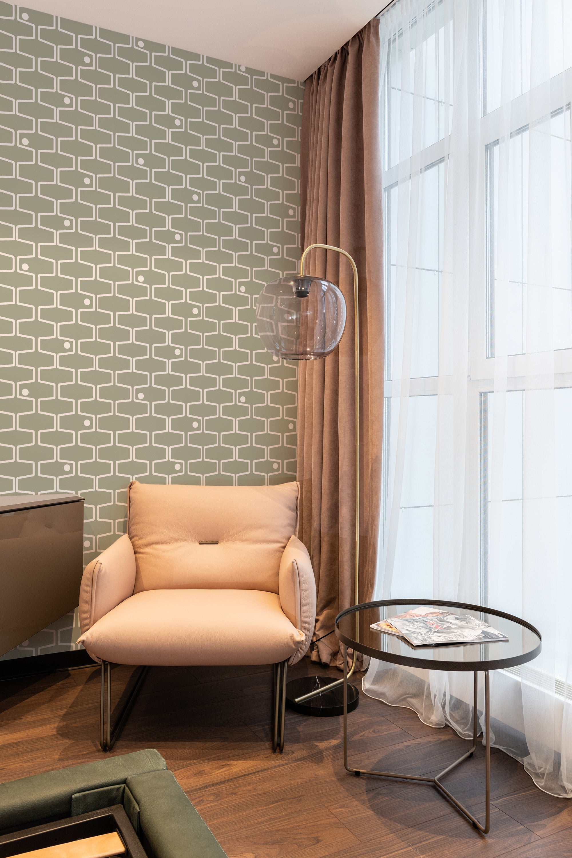 Green and white Retro geometric line wallpaper with a repeating pattern of intersecting curved lines on the wall behind a beige armchair in a cozy room, traditional wallpaper.