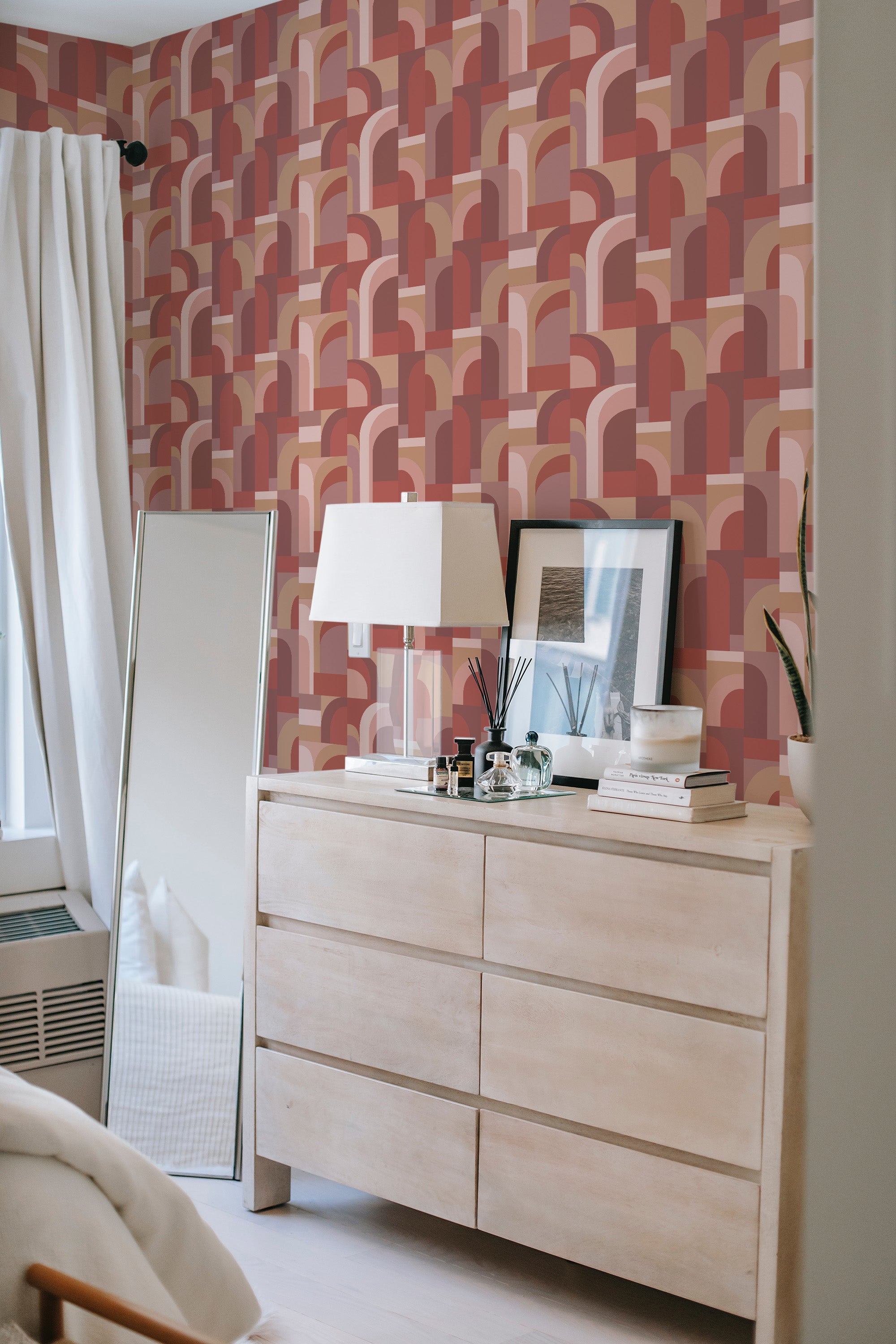 A room with Retro 70s wallpaper on the wall, displaying a pattern of bold, curved arches in shades of pink, red, orange, and beige, creating a vibrant, geometric design. A white curtain and light wooden dresser complement this traditional wallpaper.