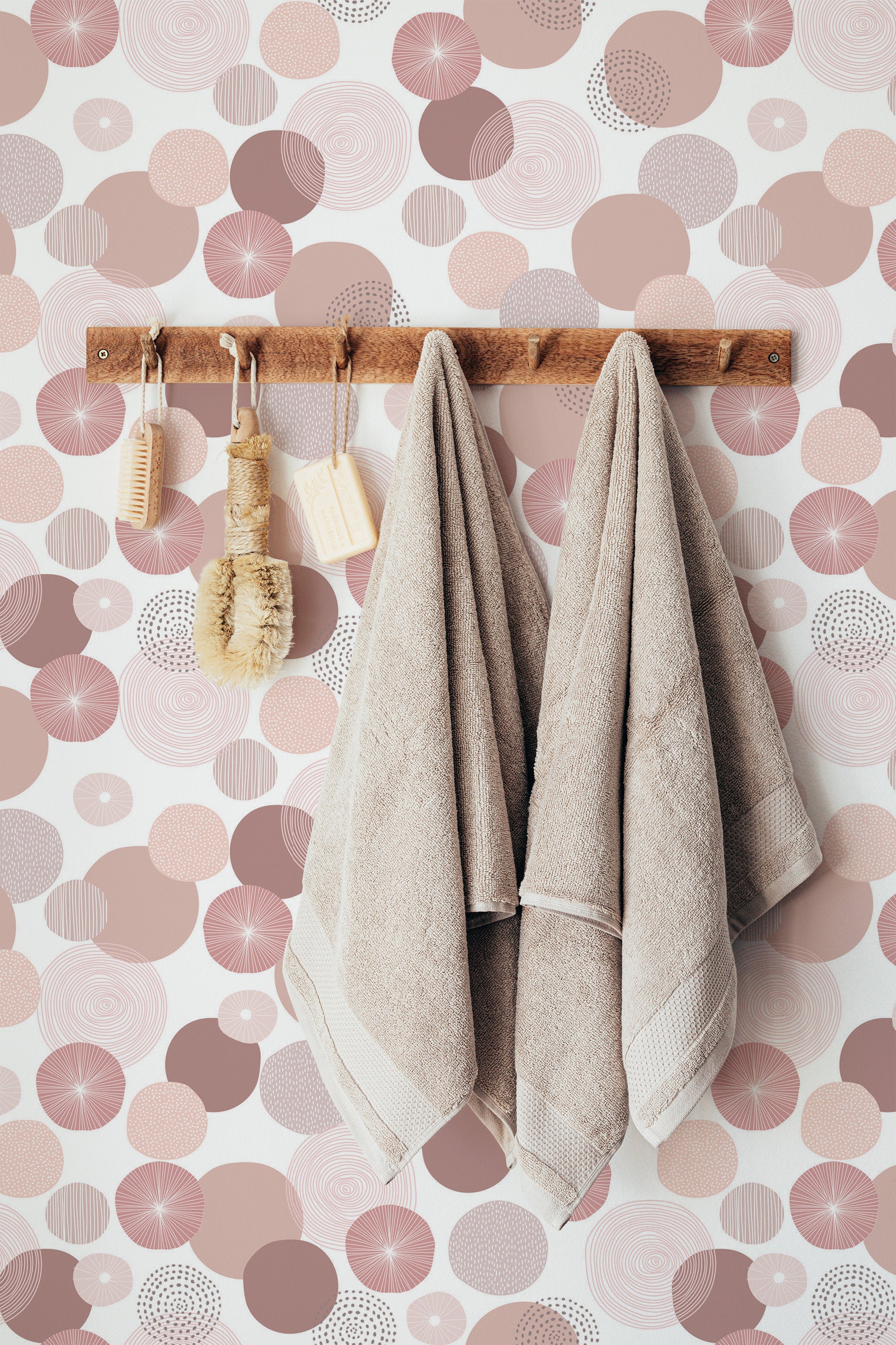 Self-adhesive wallpaper with a pink boho circle pattern in varying shades of pink, beige, and white with intricate line and dot details, behind a wooden towel rack with beige towels hanging.