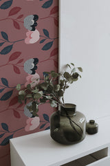 Self-adhesive wallpaper with a bold floral pattern in shades of pink, grey, and green, complemented by green leaves, applied on a room's wall, with a plant in a green vase on a white shelf in the foreground.