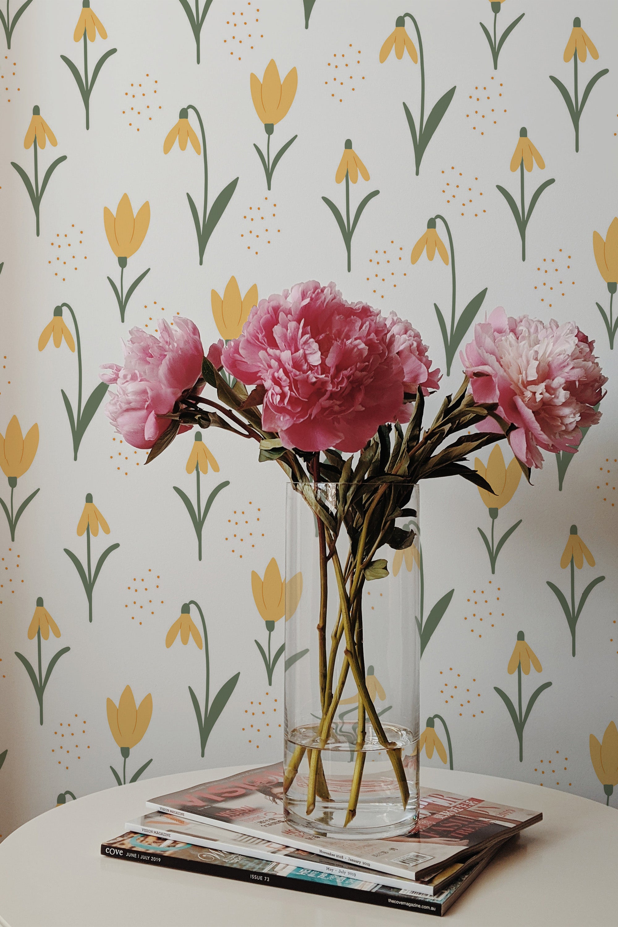 Self-adhesive wallpaper with a pattern of yellow tulips and green stems on a pale background, accented with a vase of pink peonies on a table.