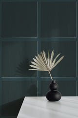Self-adhesive dark green geometric-patterned wallpaper on a room wall with a decorative vase and dried plant on a white surface.