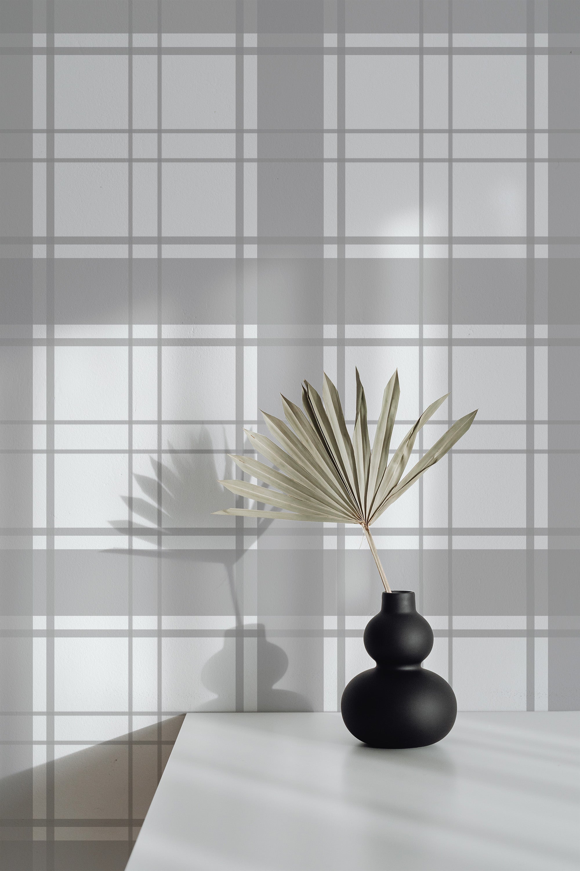 Self-adhesive plaid square pattern wallpaper in white and gray tones with shadows cast on it, accented by a small black vase with a dried palm leaf on a white table.
