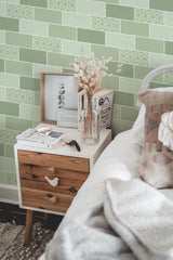 Self-adhesive green tile-patterned wallpaper with a geometric design on a bedroom wall, complementing a wooden bedside table and cozy bedding.