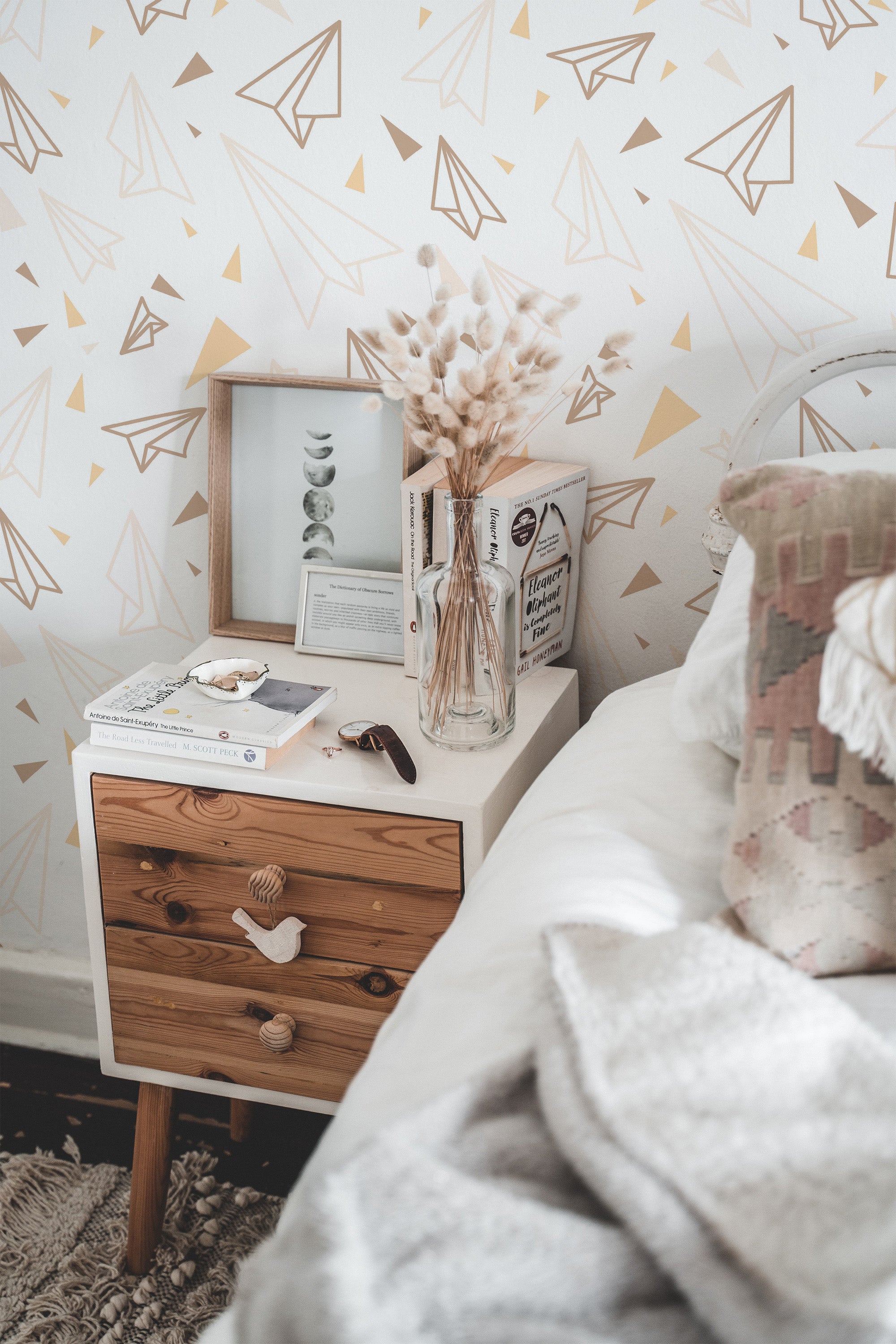 Self-adhesive wallpaper with a geometric paper plane pattern in white and gold on an off-white background, in a cozy room with a bedside table and decor.