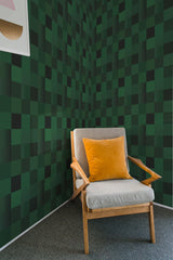 A room with a wall covered in dark green tiled pattern with varying shades creating a 3D illusion, accompanied by a wooden armchair with a yellow cushion, embodying traditional wallpaper.