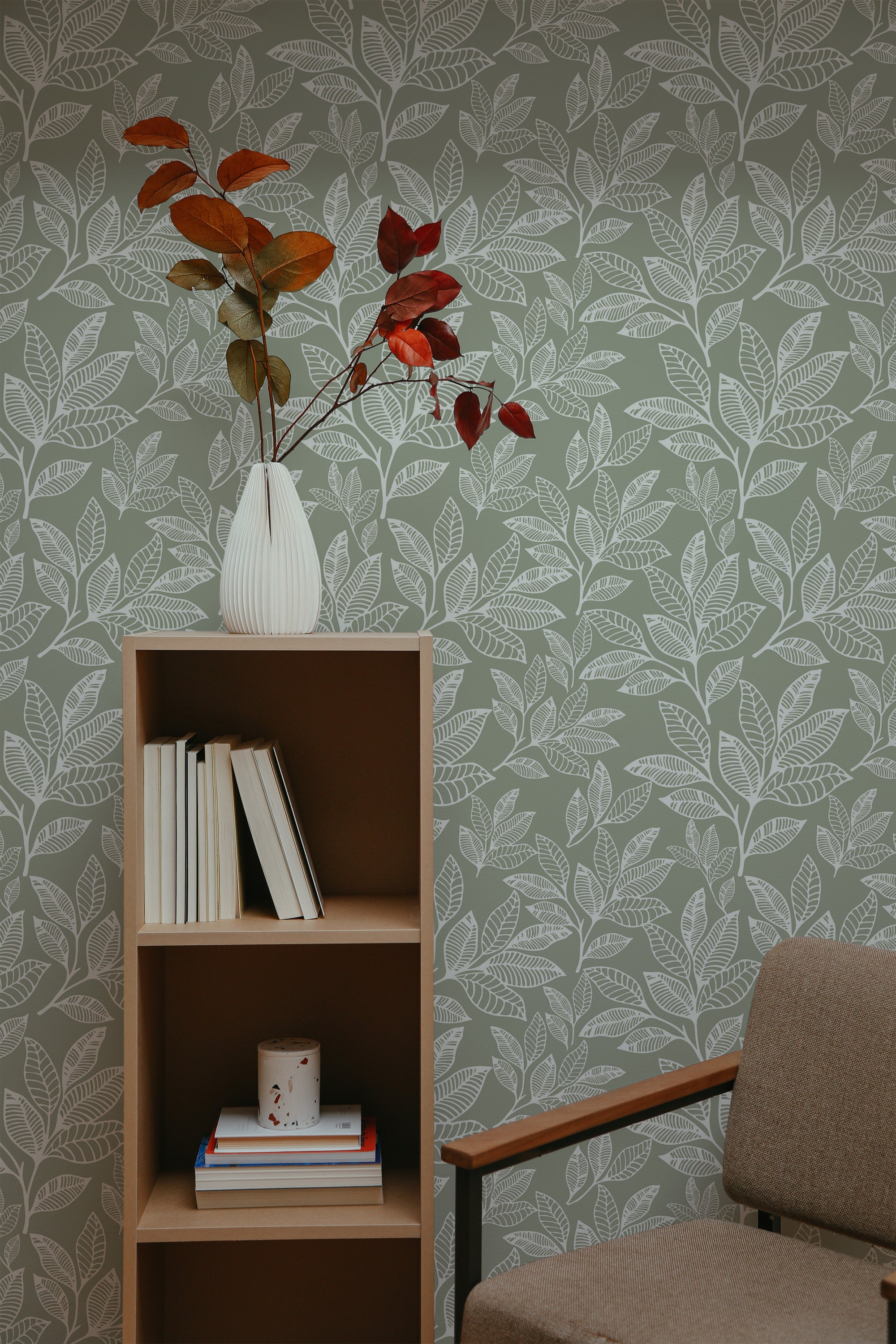 Self-adhesive leaf line art wallpaper with a pattern of stylized leaves in varying shades of green, creating a natural and calming backdrop in a room with a wooden shelf and a cozy armchair.