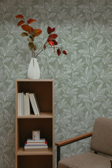 Self-adhesive leaf line art wallpaper with a pattern of stylized leaves in varying shades of green, creating a natural and calming backdrop in a room with a wooden shelf and a cozy armchair.