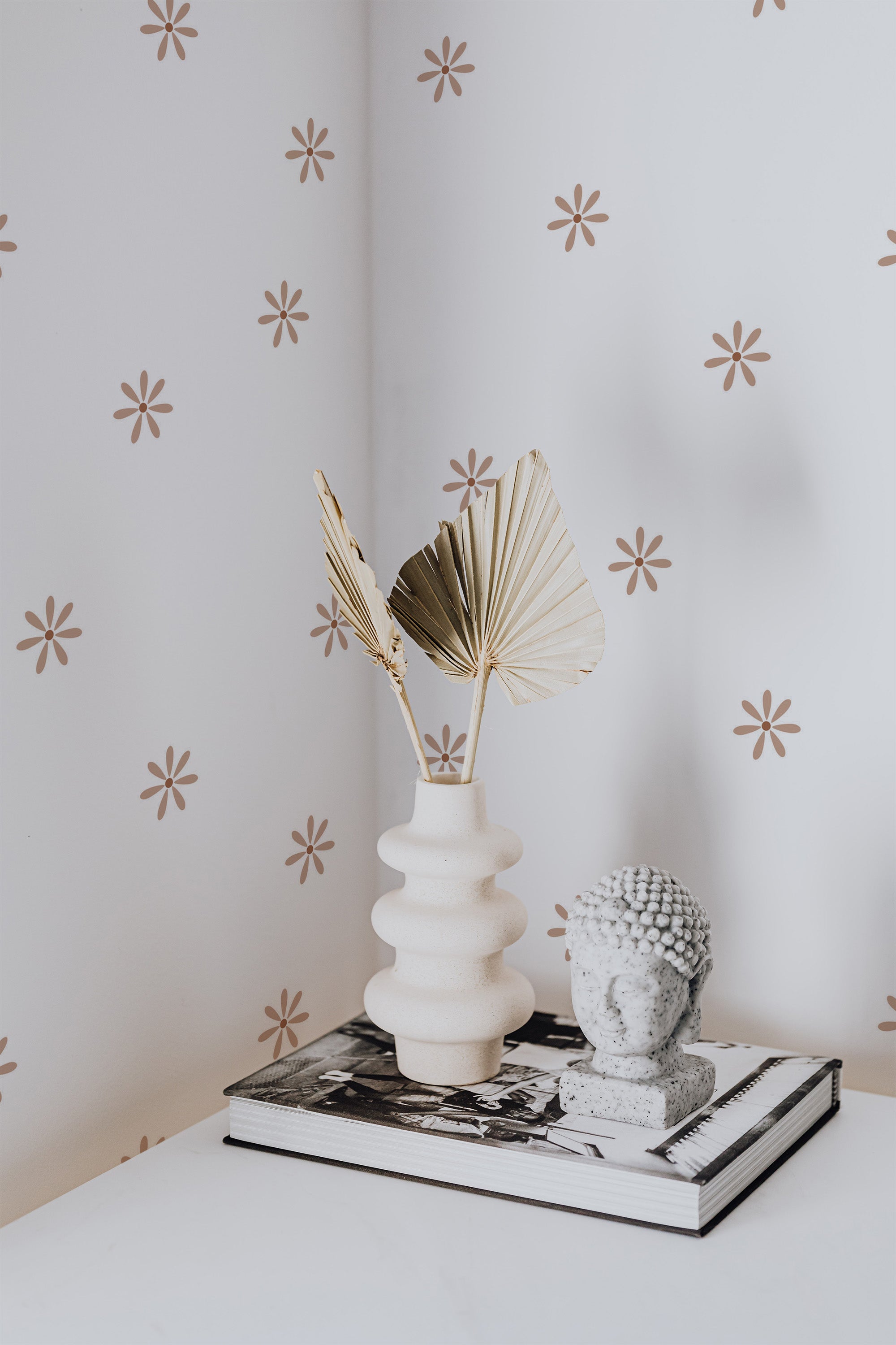 Self-adhesive beige minimal daisy pattern wallpaper on a wall with a decorative vase, books, and ornamental piece on a table in the foreground.
