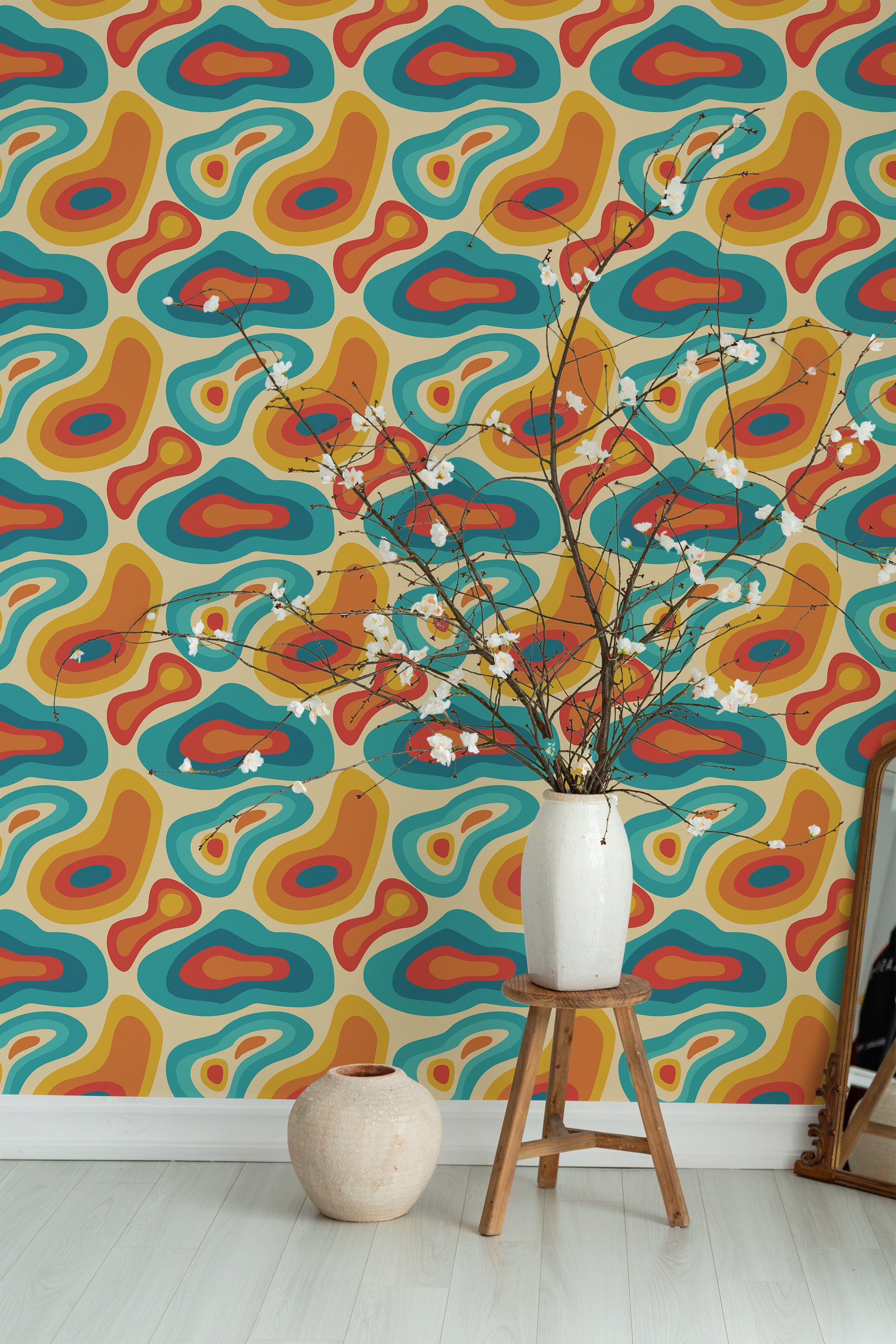 Self-adhesive wallpaper with a colorful retro pattern of abstract shapes in blue, orange, and brown tones on a room wall, complemented by a vase on a wooden stool.