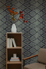 Self-adhesive dark snake skin patterned wallpaper with geometric scales in a room, accompanied by a wooden bookshelf, a vase with red leaves, and a brown armchair.