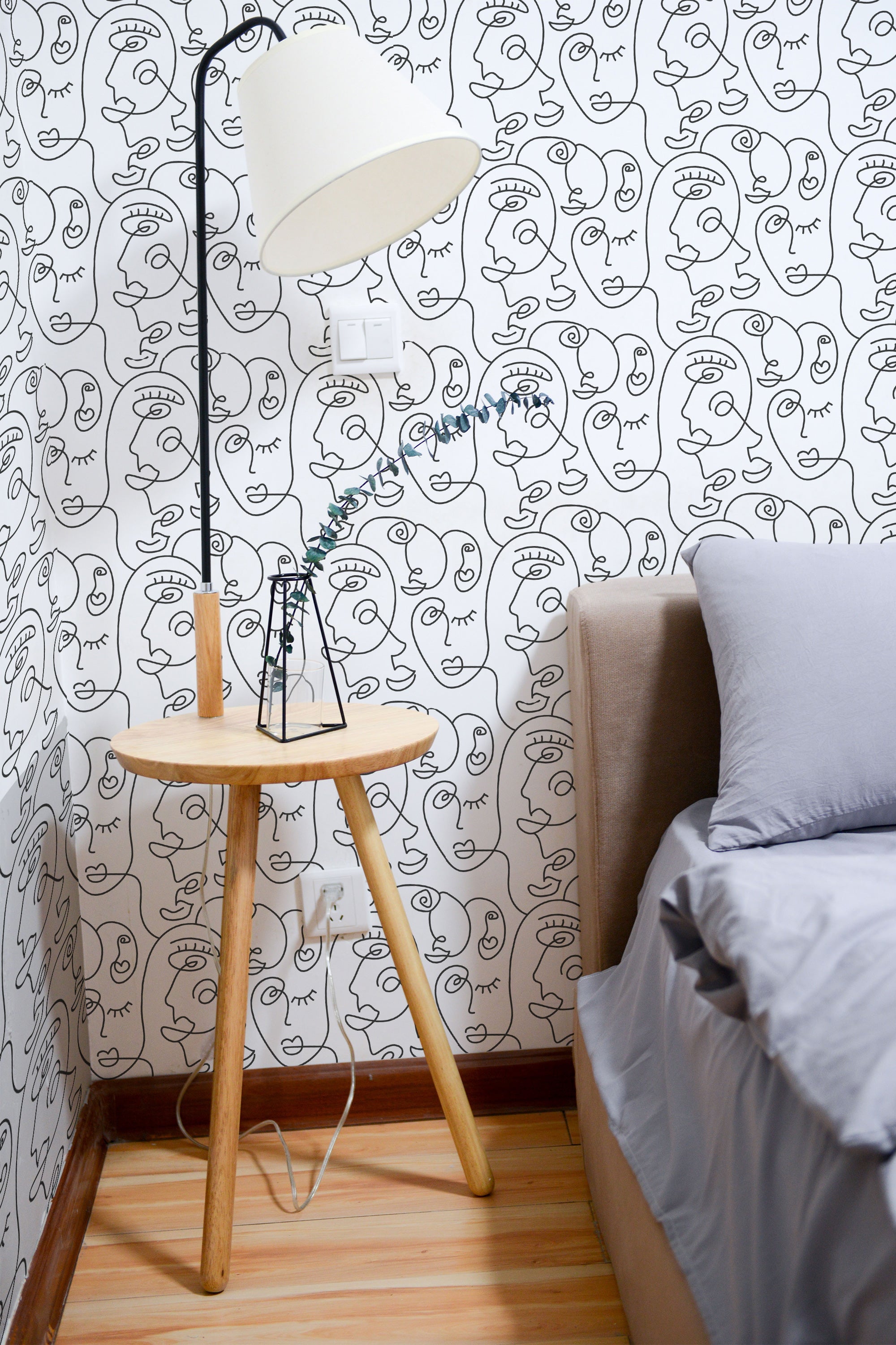 Self-adhesive wallpaper with continuous line drawings of abstract faces in black on a pale background, covering a bedroom wall.