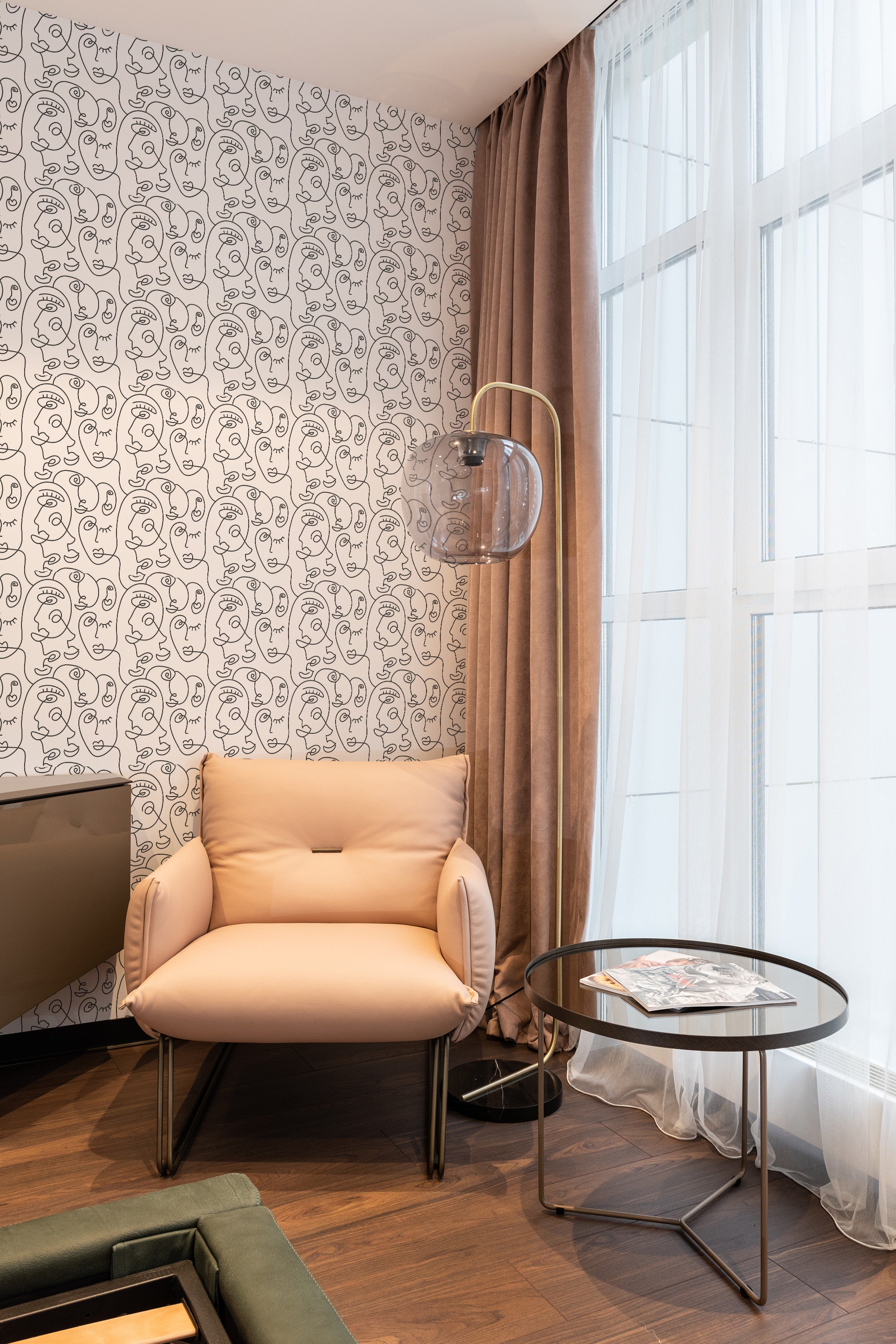 A room with walls covered in face line art wallpaper, illustrating continuous abstract facial profiles in black on a white background, complemented by a cream armchair, sheer curtains, and traditional wallpaper.