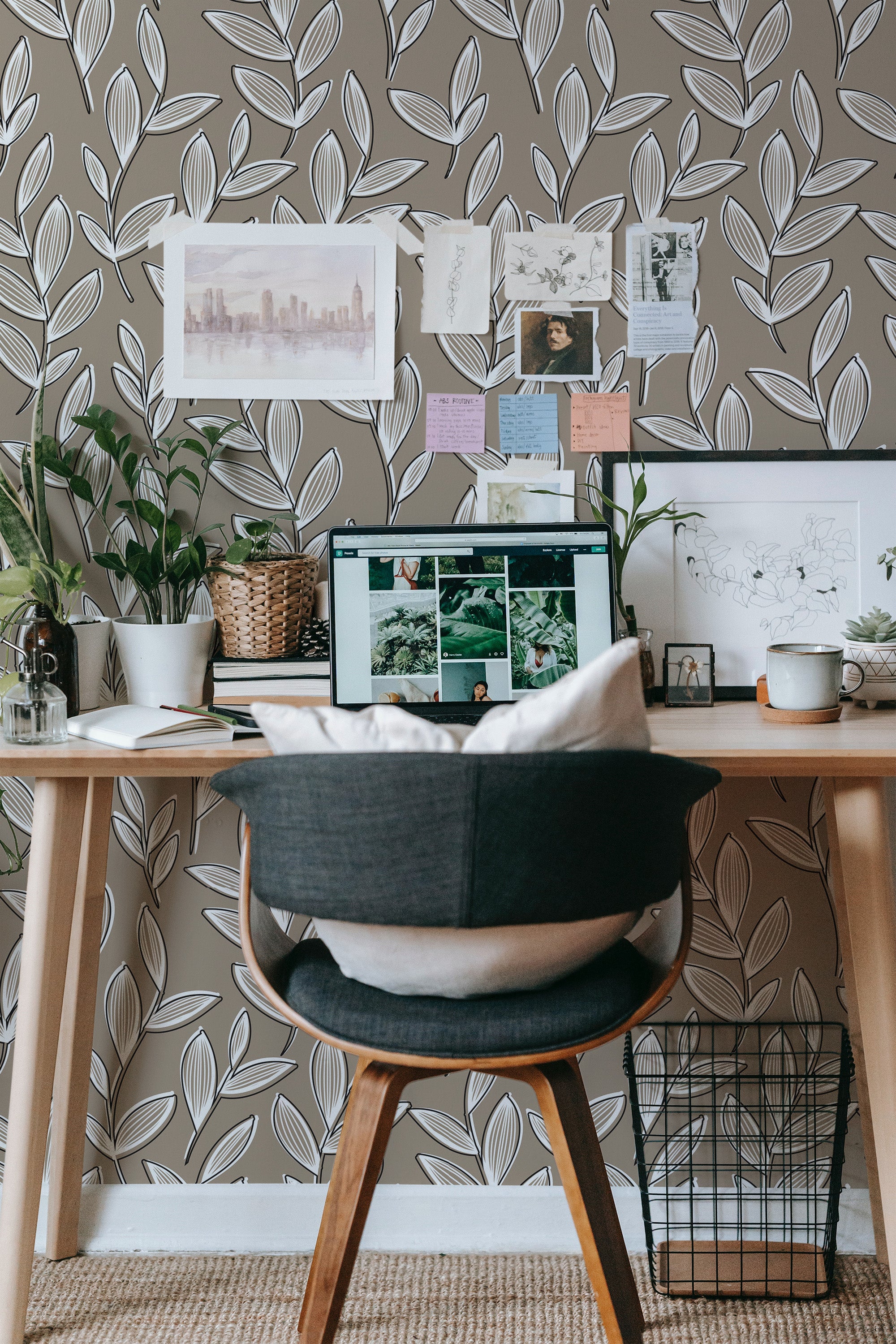 Self-adhesive wallpaper with a neutral leaf pattern on a light background, featuring white and gray overlapping leaves, covering the wall behind a wooden desk adorned with indoor plants, art prints, and a computer monitor.