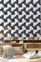 An image of a room where the wall has a backsplash wallpaper design with a bold, geometric black and white checkerboard pattern, creating a striking visual effect. The floor is covered with a light-colored rug, and natural woven furniture, including a rattan chair and wicker baskets, adds a warm, organic contrast to the modern wall design. Potted plants bring a touch of greenery, blending contemporary with earthy tones in a room adorned with traditional wallpaper.
