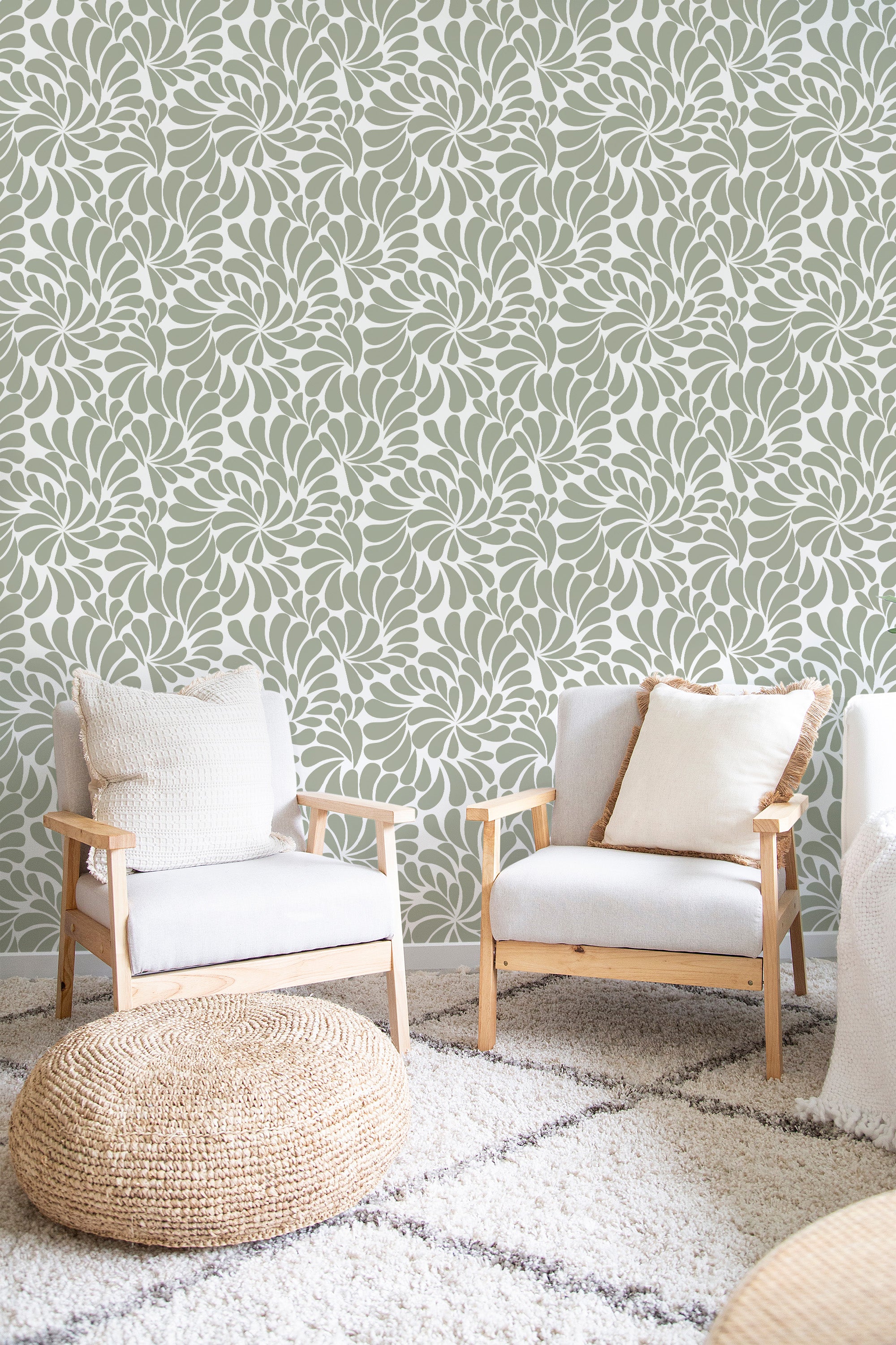 A room with green Paisley design wallpaper that has intricate, swirling patterns creating a floral-inspired look. The room contains two wooden chairs with cream cushions and a round woven ottoman, all on a soft rug. Traditional wallpaper.