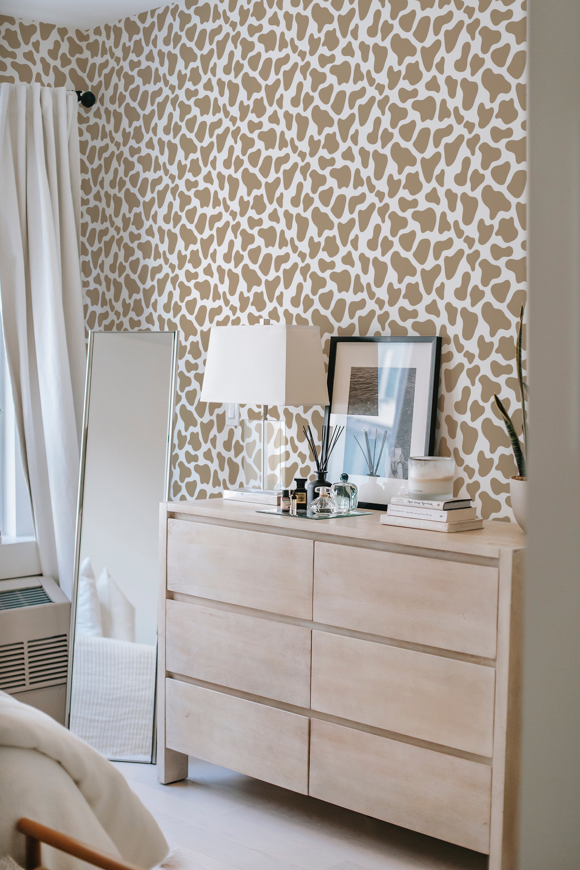 The wall is covered in a giraffe-like pattern with irregular shapes in beige and off-white tones. The room contains a light wooden dresser, a lamp with a white shade, a framed picture, and a mirror leaning against the wall. Traditional wallpaper.