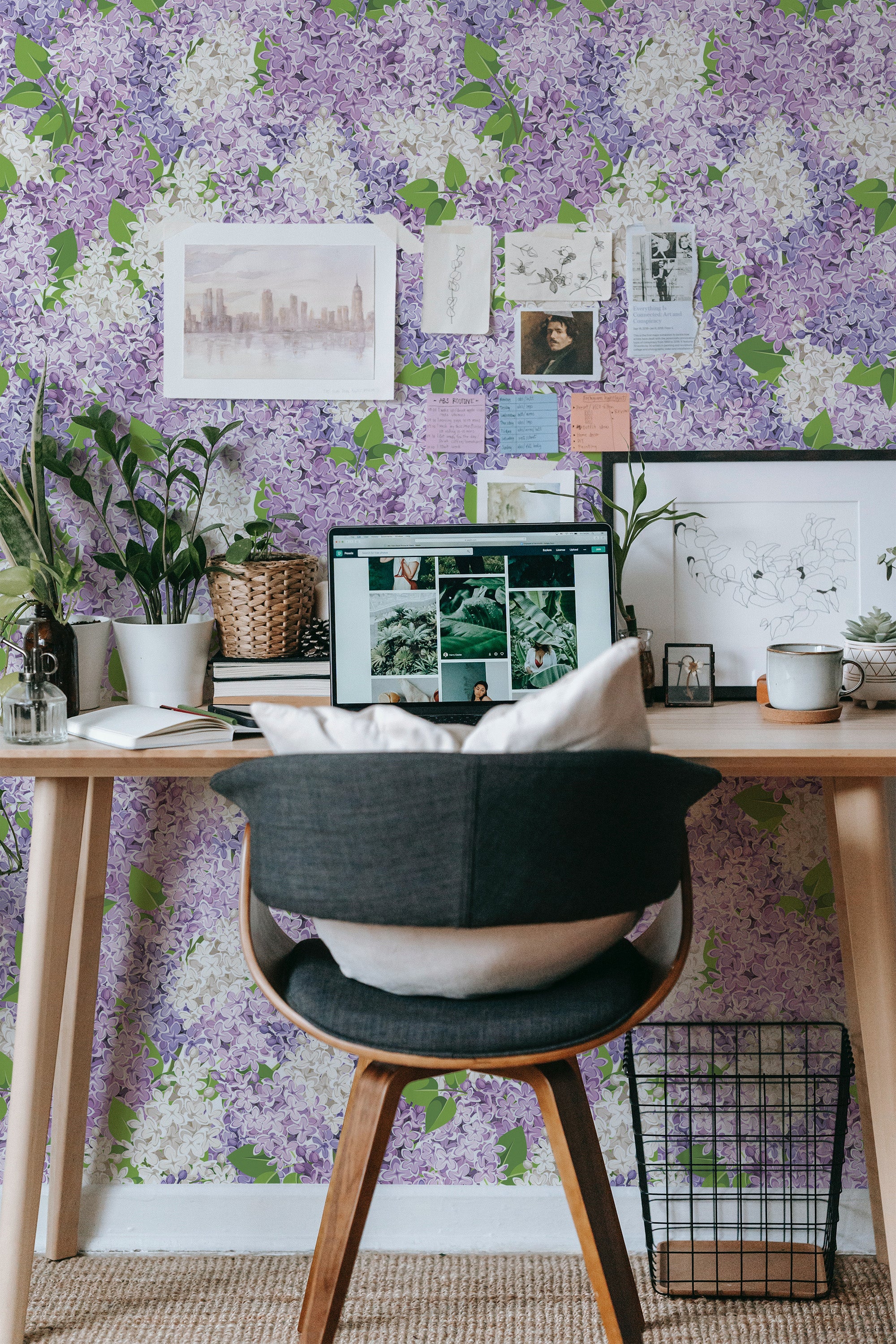 Self-adhesive lilac wallpaper with dense purple lilac clusters, green leaves, and white accents creates a vibrant and lively atmosphere on the wall behind a wooden desk. The floral pattern adds a touch of freshness to the room, complemented by plants and wall art.