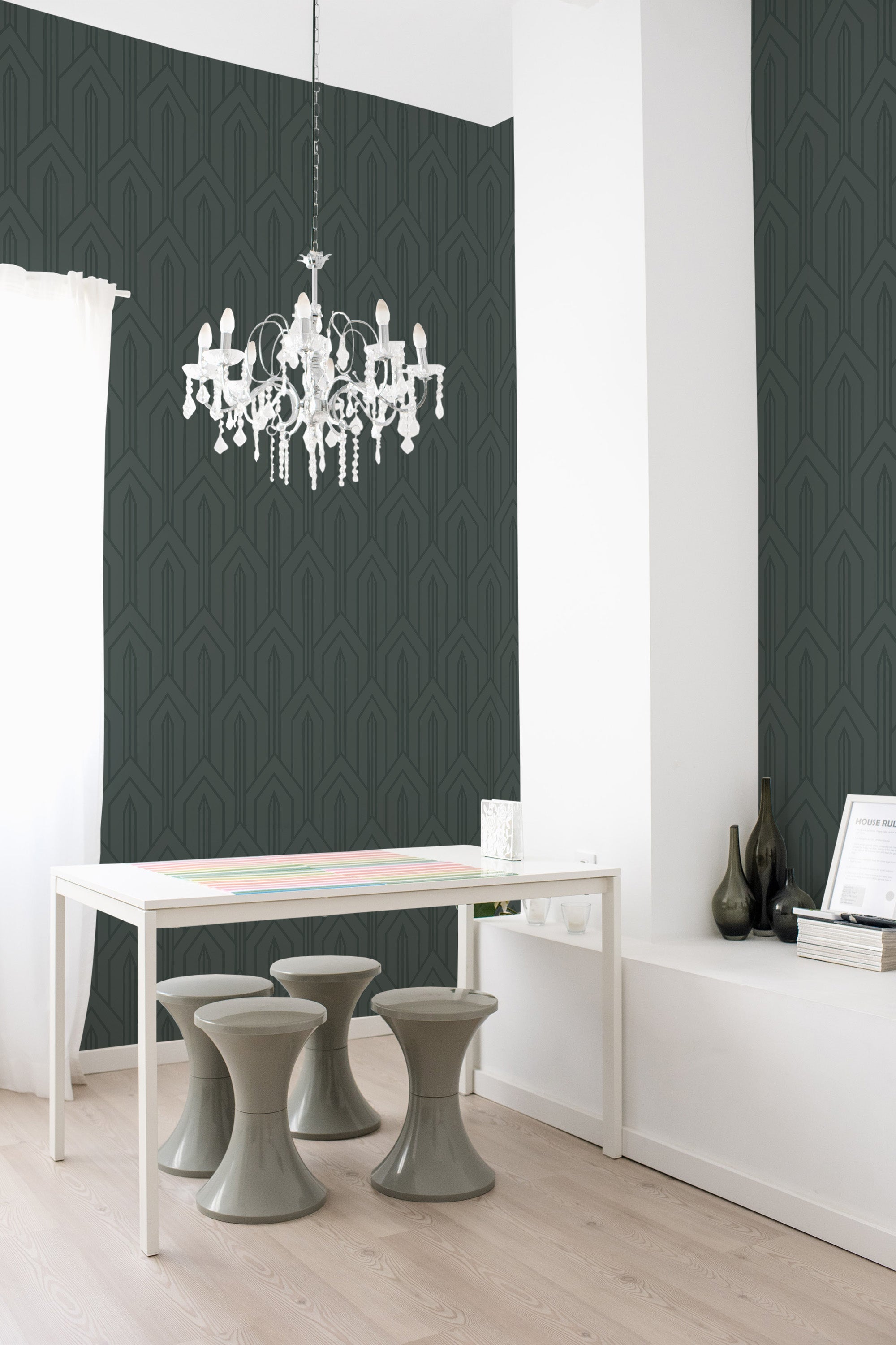 A room with a dark art deco wallpaper with a geometric pattern of repeating arches and angular lines in a muted green tone, complemented by a simple white desk and modern gray stools under an ornate chandelier, traditional wallpaper.