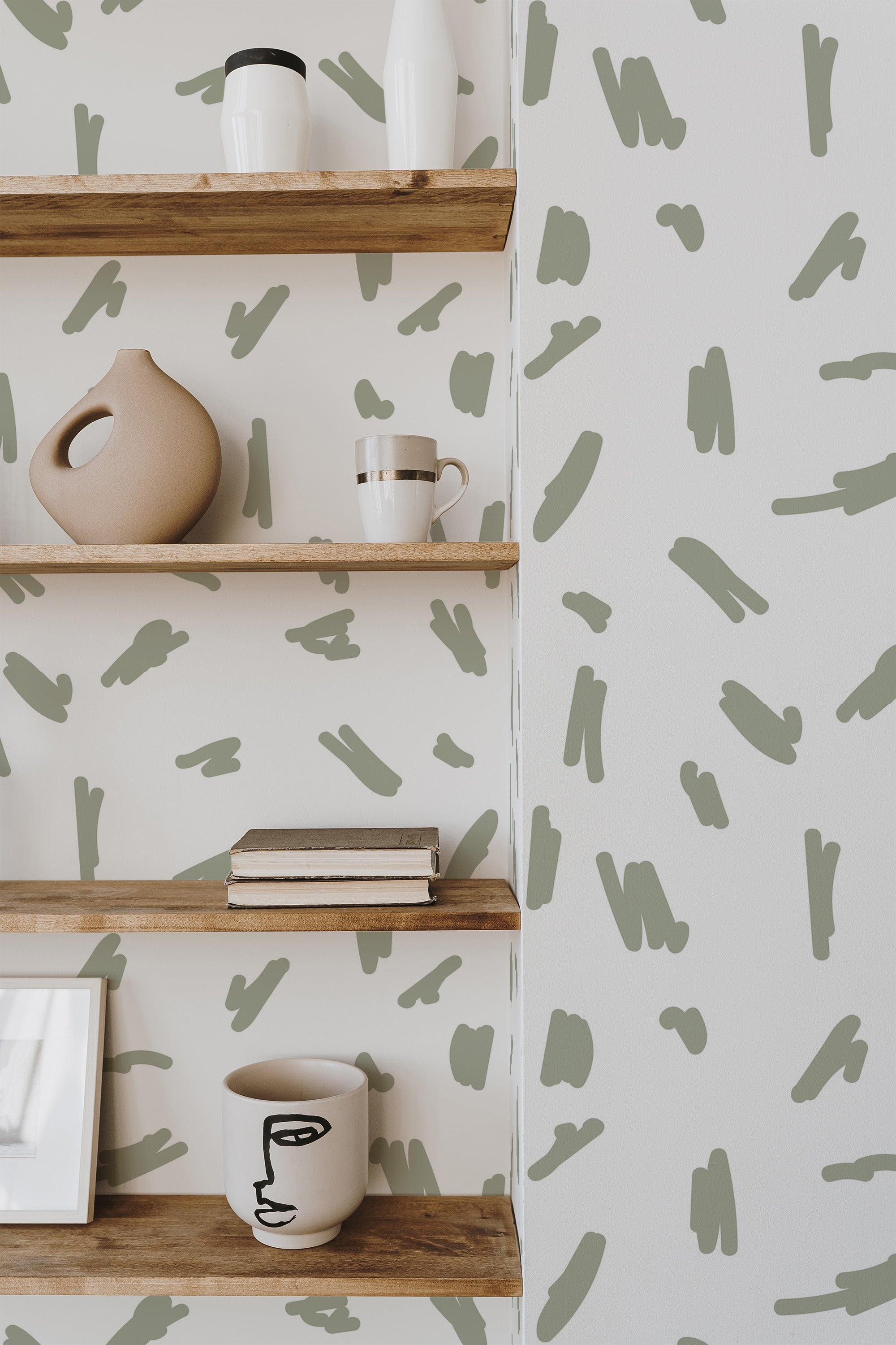 Self-adhesive wallpaper with abstract brush stroke design in olive green on a soft white background, complemented by minimalist wooden shelves with vases and a coffee mug.