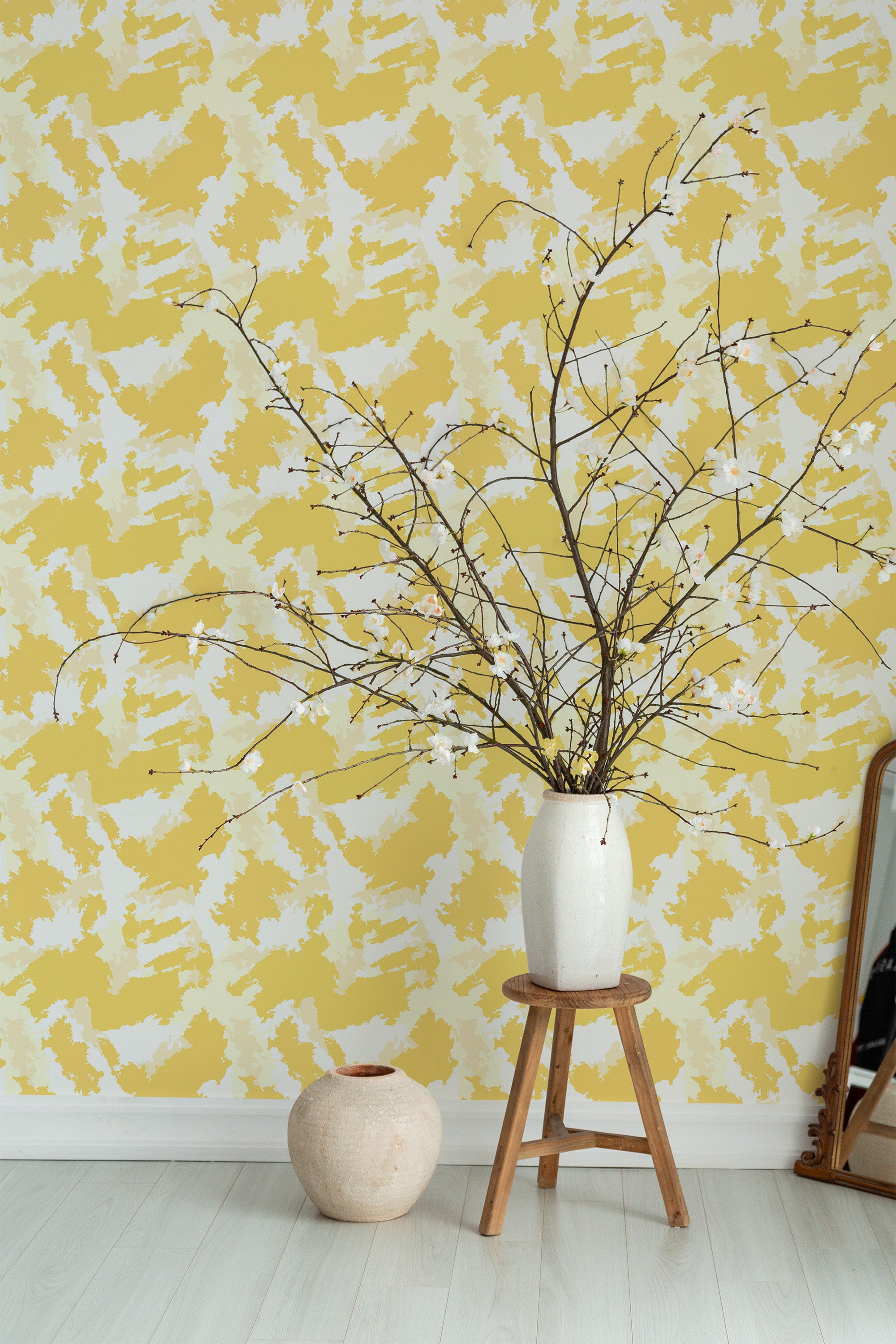 Self-adhesive wallpaper with a yellow watercolor pattern, creating an abstract brushstroke effect on the wall. The room includes a simple wooden stool, a white vase with bare branches, and a rustic round vase on the floor in front of a large mirror.