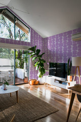 A living room with purple retro shape pattern wallpaper on the wall, consisting of a repeated design of irregular rectangular blocks in soft, varying shades of purple. The wallpaper adds a vintage and bold aesthetic to the vibrant, well-lit room, enhancing the modern decor and natural light. Traditional wallpaper.