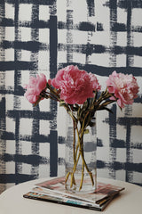 Self-adhesive wallpaper with a Navy blue brush stroke design on a light background, creating a modern and dynamic grid pattern. Pink flowers in a clear vase are placed in front of the wallpaper, adding a touch of softness to the room's d��cor.