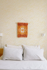 Self-adhesive wallpaper with a tiny speckled dot design in yellow on a cream background, covering the entire wall behind a bed with white pillows and linens.