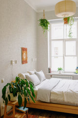 A room with light gray wallpaper resembling a subtle marble pattern, adding a soft, elegant texture to the walls. The wallpaper complements the neutral-toned bedding and natural wooden furniture, creating a calm and cozy atmosphere. Hanging plants, soft lighting, and a large window inviting natural light enhance the serene ambiance. The design choice reflects a classic and timeless aesthetic, fitting for traditional wallpaper.